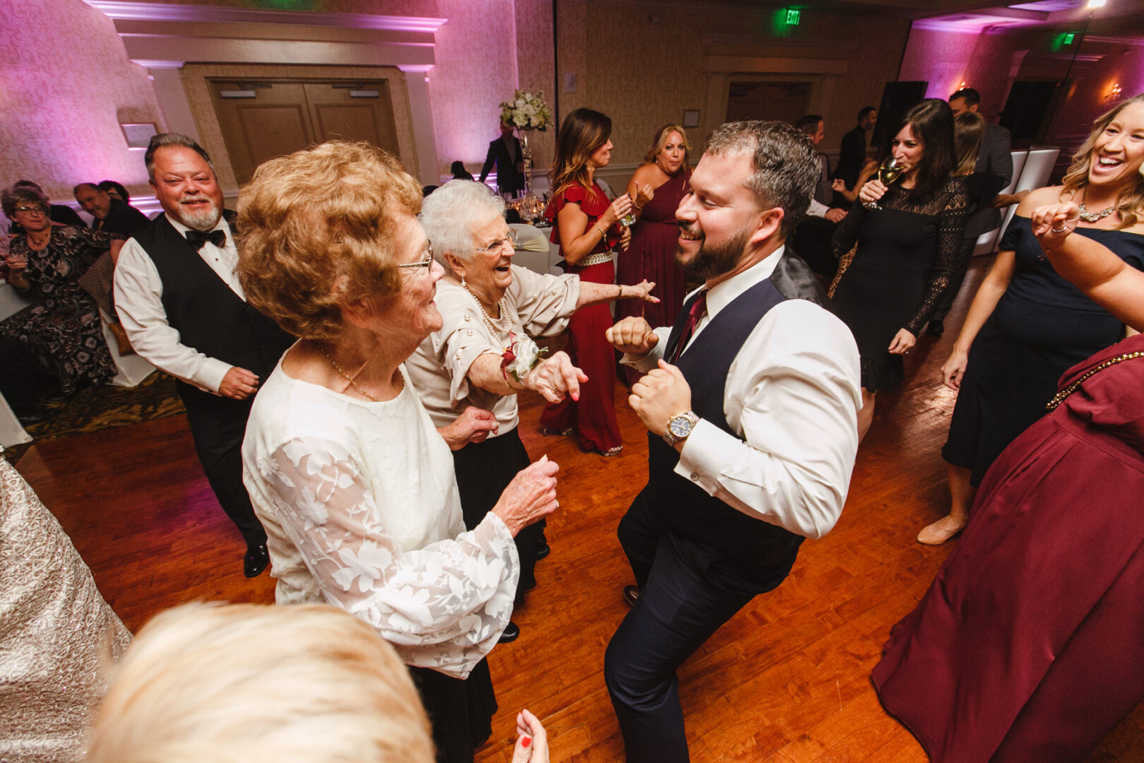 Guests of various ages are dancing energetically on the polished wooden floor of the Windsor Ballroom, their elegant attire flowing with every move at this formal wedding event.