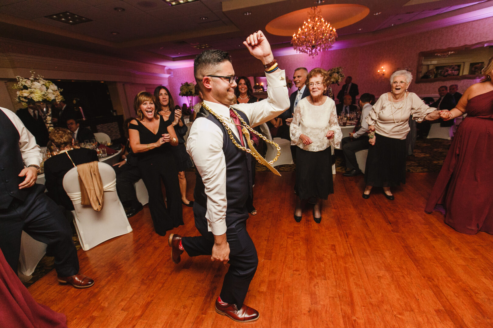 A man in formal attire dances energetically on the wooden floor of the Windsor Ballroom, surrounded by cheering and clapping guests at a lively wedding celebration.