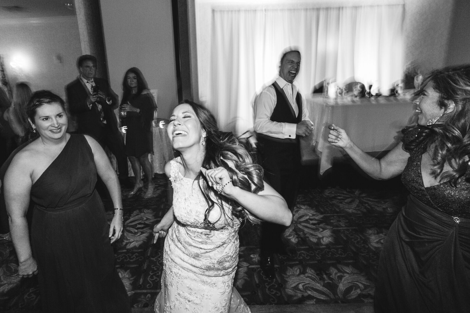 People dancing joyfully at an indoor event, with a bride in the center wearing a white dress. The atmosphere is lively with smiles and movement at the Windsor Ballroom Wedding, all captured in an elegant black and white setting.