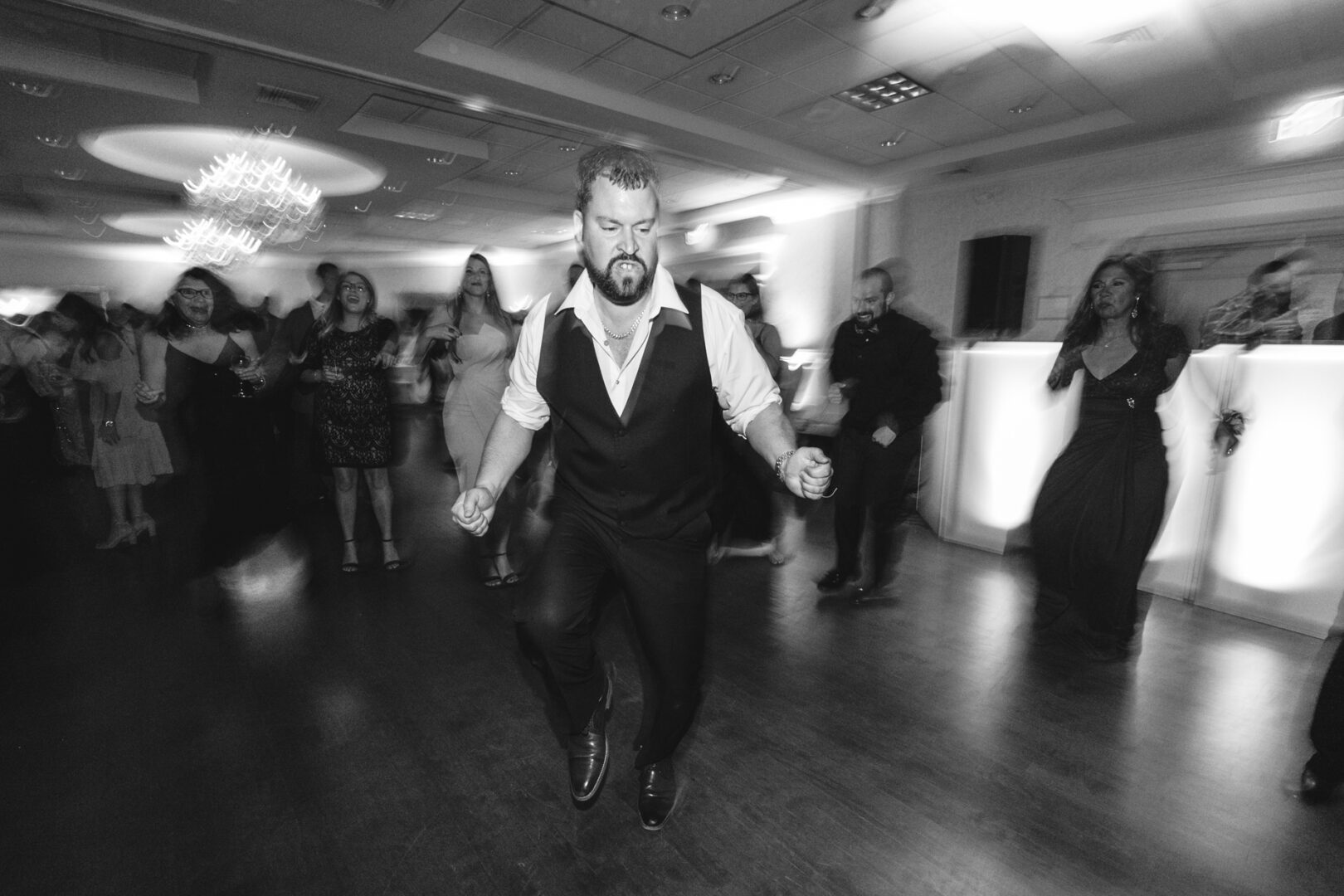 In the elegant Windsor Ballroom, a man in a vest energetically dances at a wedding, surrounded by guests in the dimly lit room.