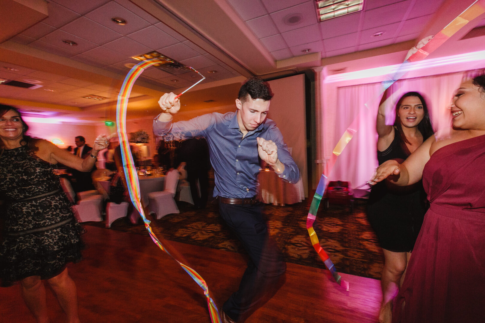 In the Windsor Ballroom, a man in a blue shirt enthusiastically waves rainbow streamers while dancing among guests in a festive, warmly lit room.