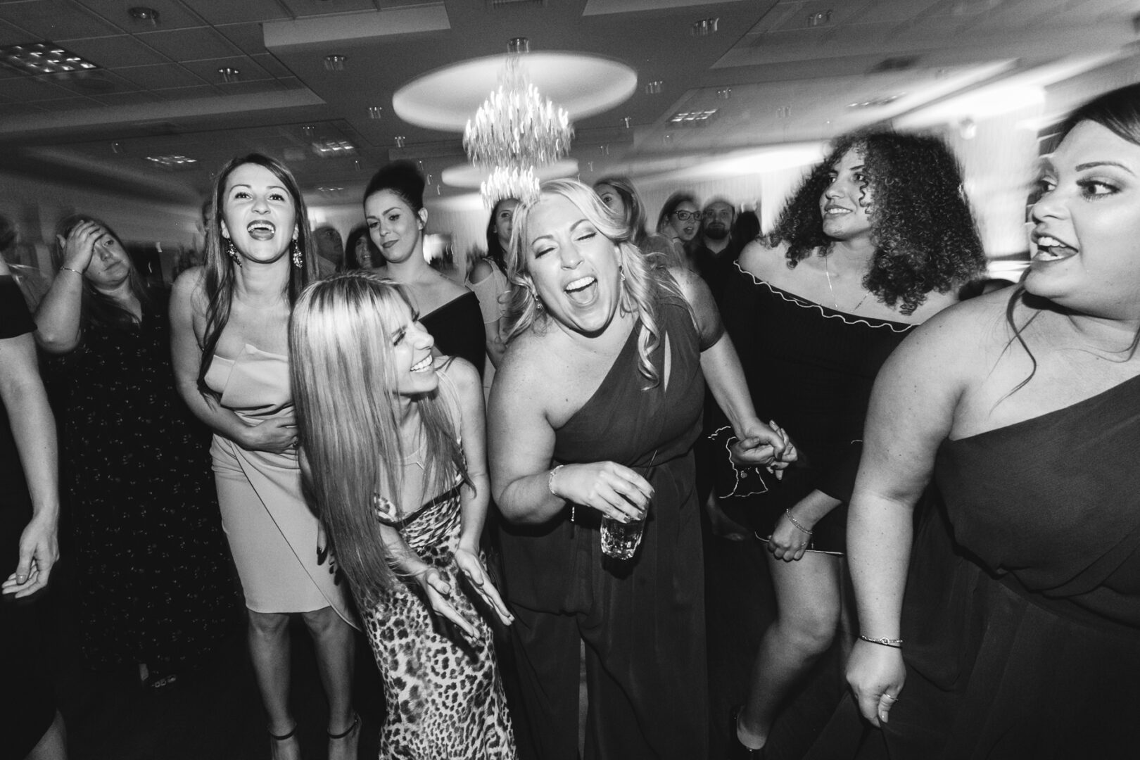 A group of people in formal attire are laughing and dancing together in the well-lit Windsor Ballroom, adding a touch of elegance to the lively wedding celebration.