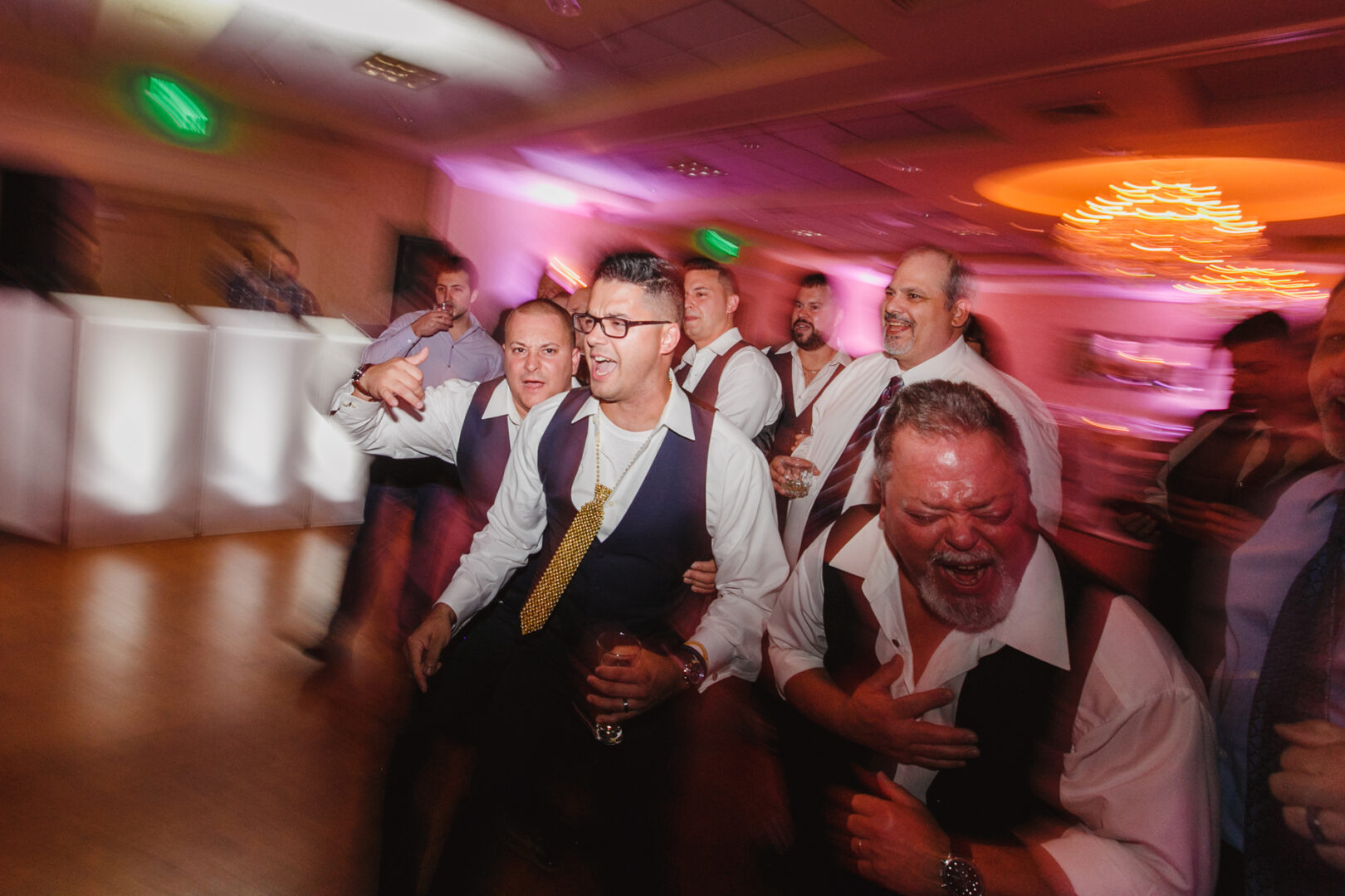 A group of men in formal attire, laughing and walking closely together in the warmly lit Windsor Ballroom, captures the joyous essence of a wedding event.