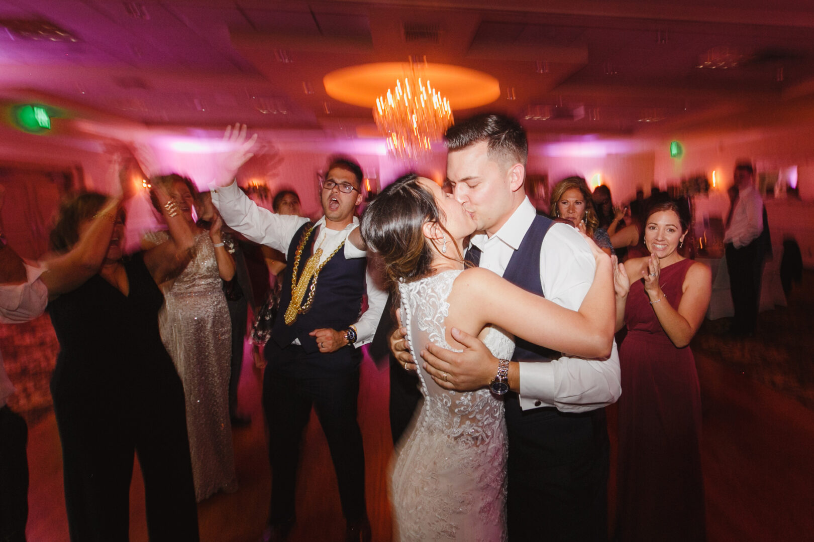 At the Windsor Ballroom wedding, a bride and groom share a kiss on the dance floor, surrounded by guests in formal attire. Blurred lights and movement fill the space, creating a dynamic atmosphere.