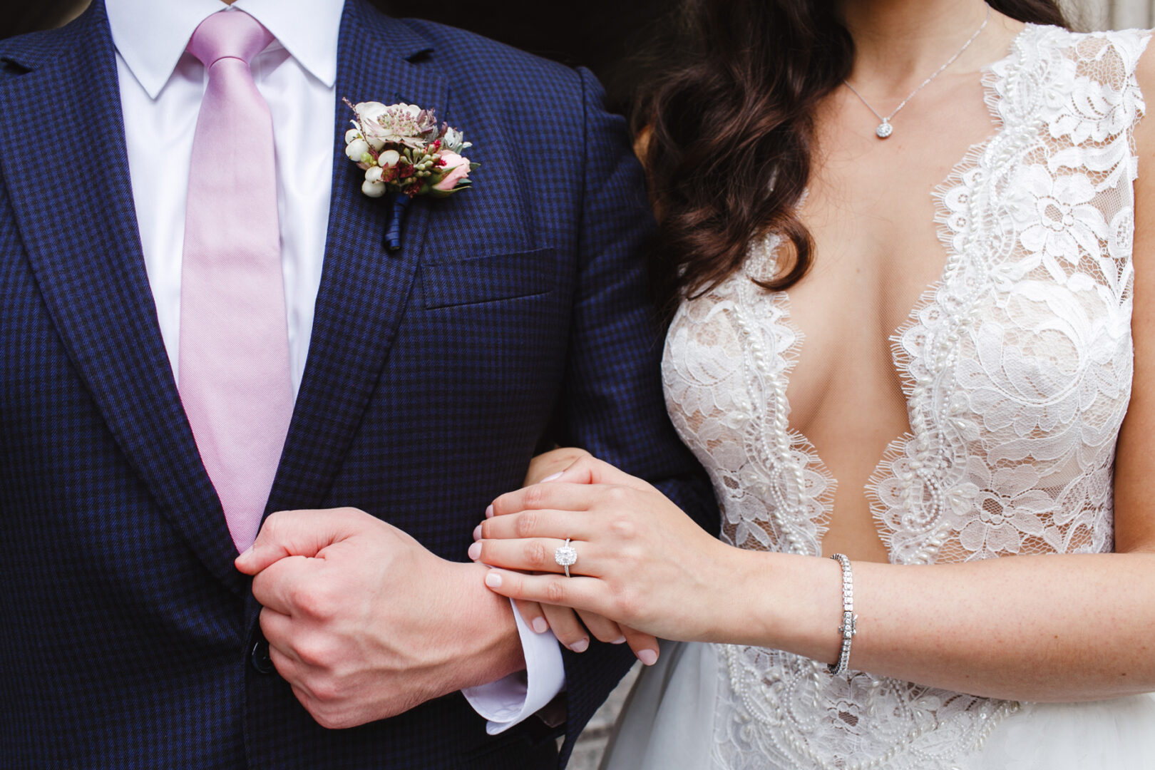 Close-up of a couple at their Chubb Conference Center wedding. The man dons a suit with a pink tie and boutonniere, while the woman, in a lace wedding dress, rests her hand on his arm, showcasing her ring and bracelet.