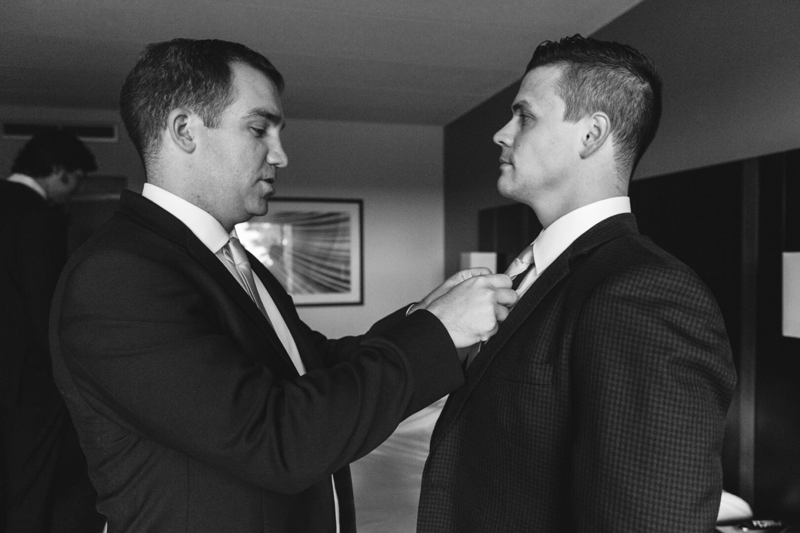 Two men in suits are preparing for a wedding at the Chubb Conference Center. One adjusts the other's tie in a room with a modern interior, setting the tone for an elegant event.