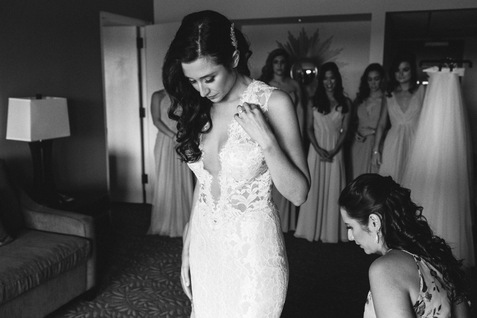 The bride adjusts her lace gown, illuminated softly in the dimly lit room of the Chubb Conference Center, while bridesmaids stand gracefully in the background.