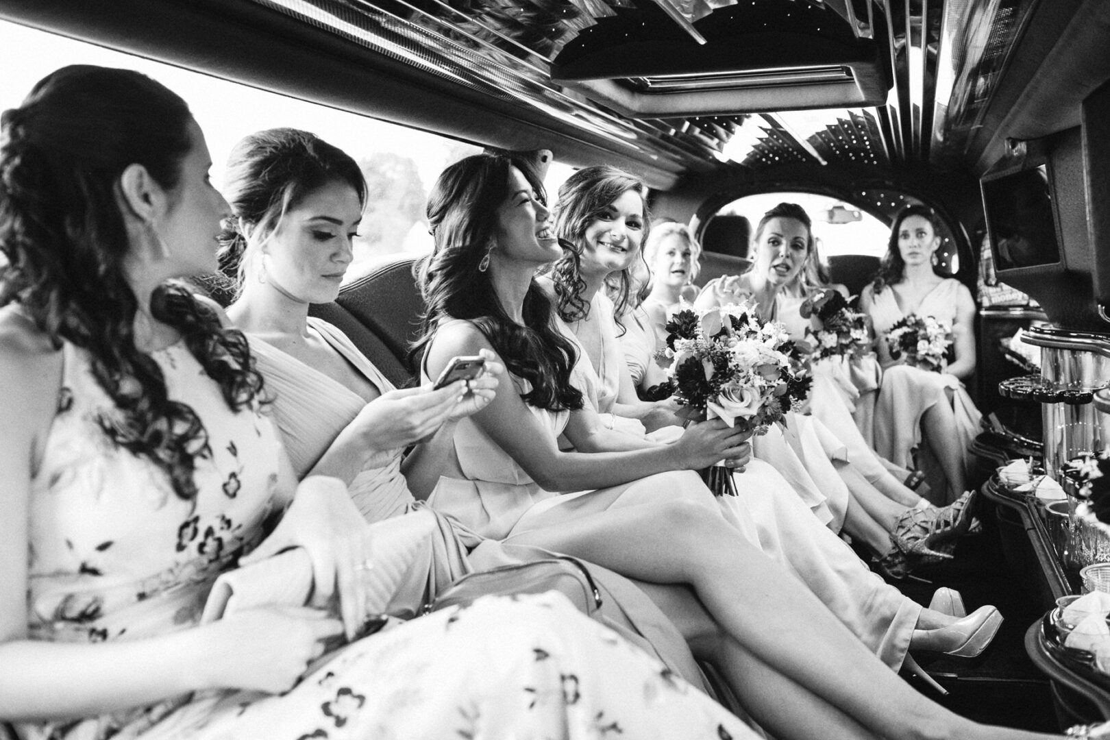 Inside a limousine, a group of women in dresses sits elegantly, bouquets in hand. This black-and-white image captures the timeless charm of a Chubb Conference Center wedding.