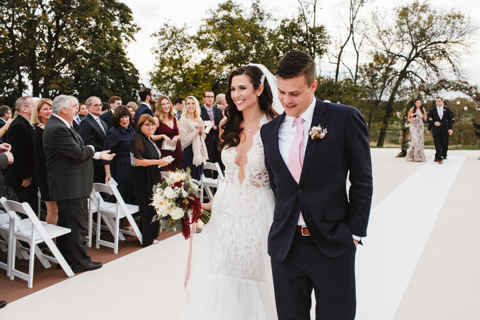 Bride and groom walk down the aisle outdoors at the Chubb Conference Center, smiling, with guests seated and standing applauding in the background.