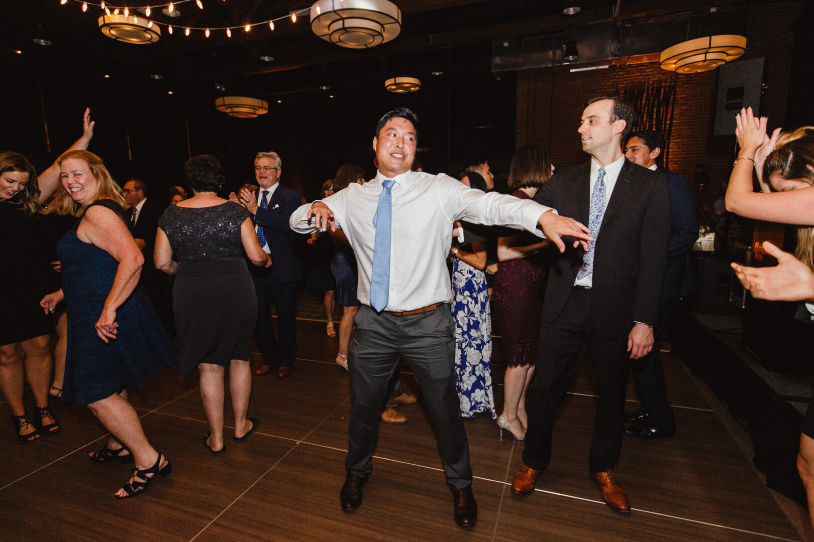 People dancing energetically in the dimly lit Chubb Conference Center, surrounded by hanging lights and brick walls, celebrating a wedding with joy.