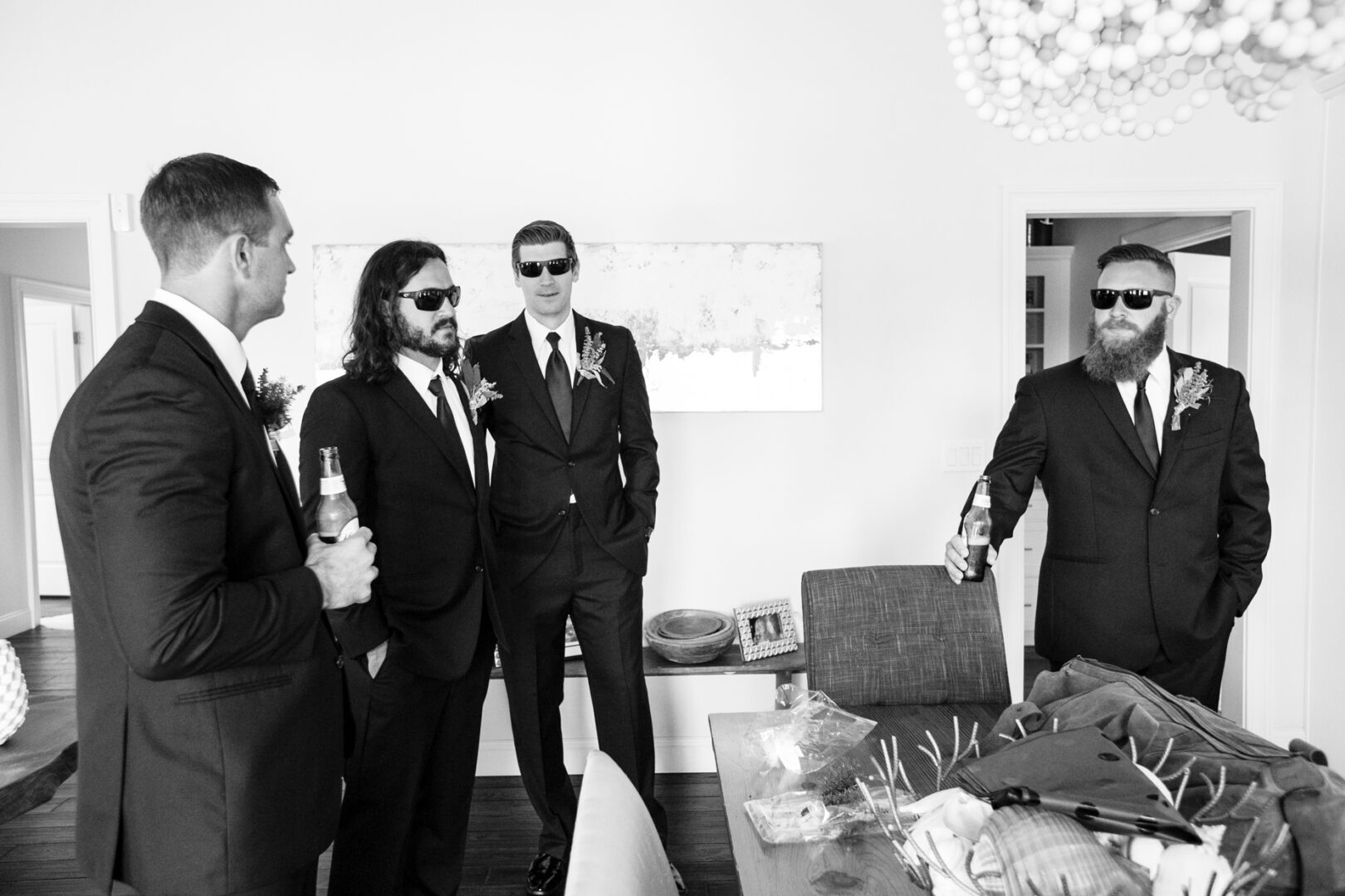Four men in suits and sunglasses stand talking at a Congress Hall wedding. Two hold bottles, while a table with various items sits in the foreground. This black and white image captures a moment of timeless elegance and style.