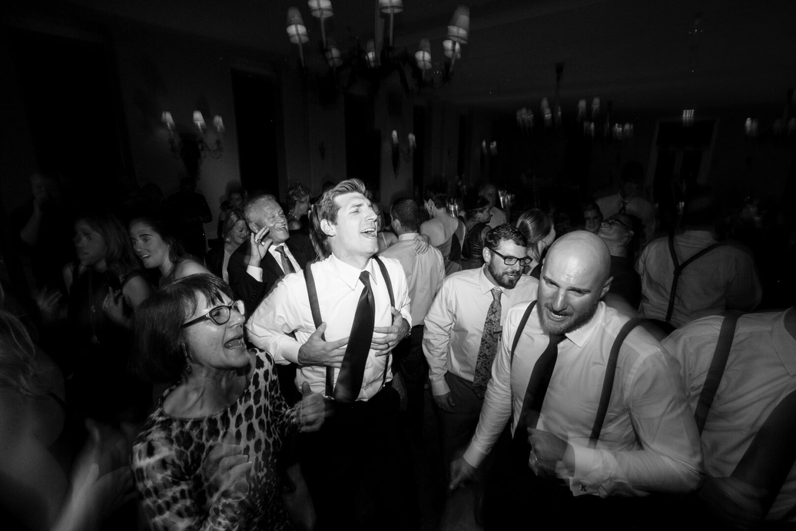 A black-and-white photo captures the lively spirit of a Congress Hall wedding, where people in formal attire and suspenders energetically dance to the music. Chandelier lights cast an elegant glow above, enhancing the joyful celebration.