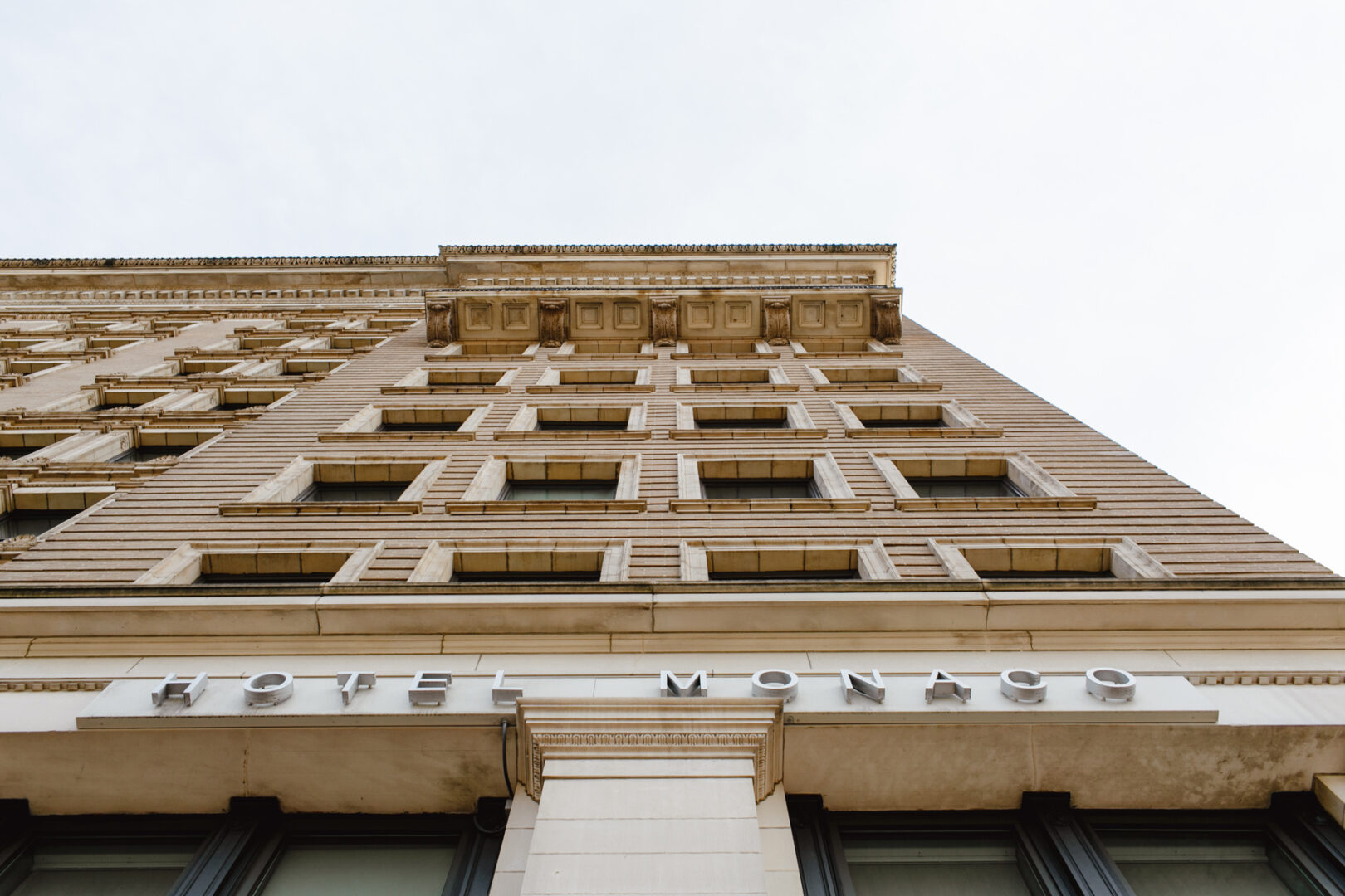 Upward view of a multi-story building with a neoclassical facade in the heart of Downtown. The words "Hotel Monaco" are displayed above the entrance, offering an elegant setting for club gatherings and sophisticated weddings.