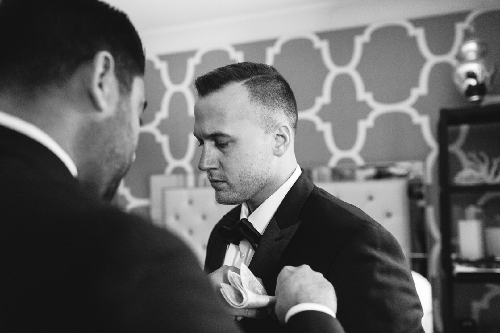 Two men in suits, one adjusting the other's lapel in a room with patterned wallpaper, shelves, and a cabinet. The black and white image evokes a classic Downtown Club vibe, as if preparing for an elegant wedding.