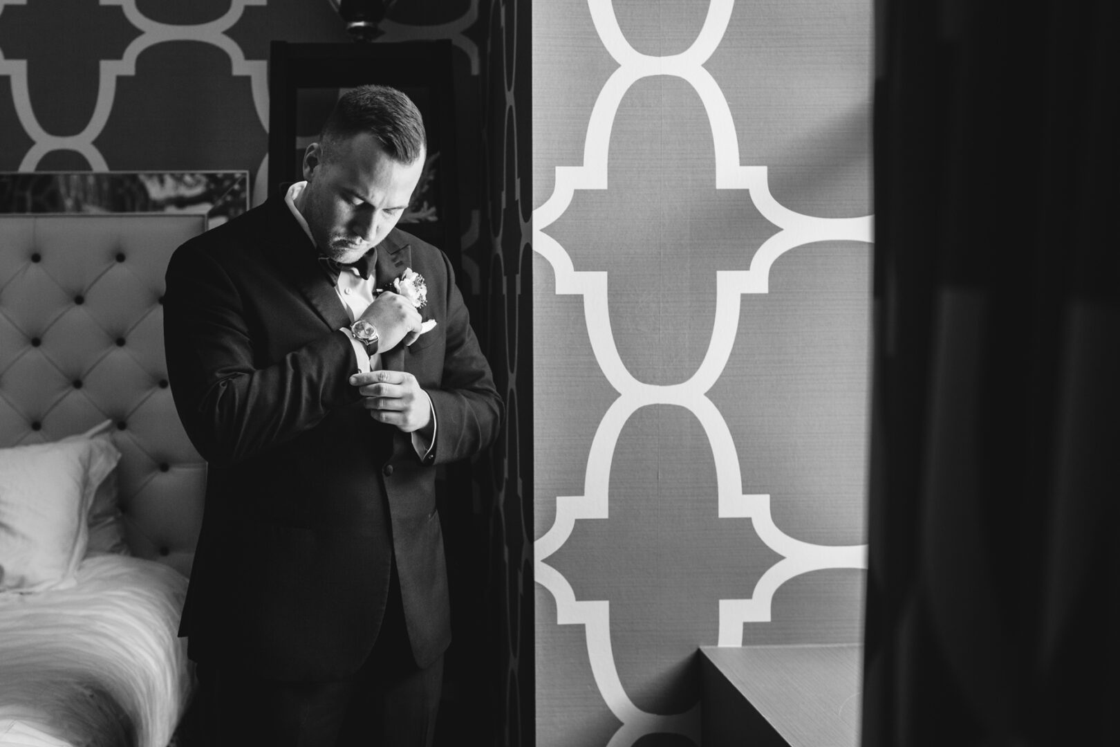 In a stylish room with a patterned wall reminiscent of a downtown club, a man in a suit adjusts his cufflink. A bed is partially visible on the left, capturing the essence of elegance and timeless charm in this black-and-white scene. .