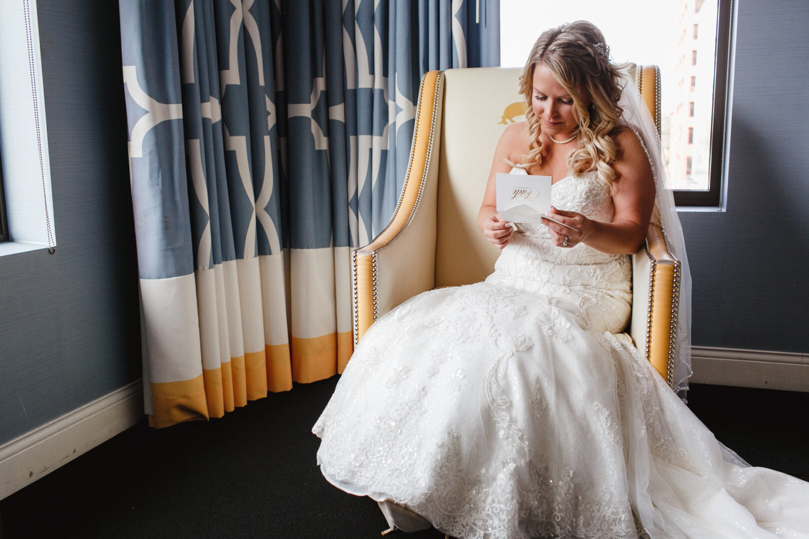 In a serene moment at the Downtown Club wedding, the bride in a white gown sits gracefully in an armchair by a window, reading a card.