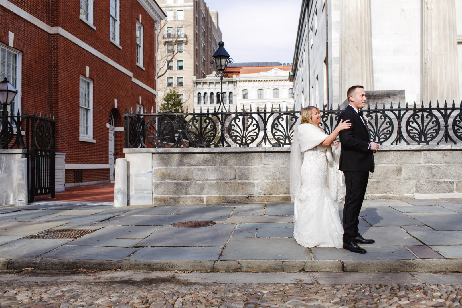 A bride in a white dress approaches a groom in a black suit from behind on the stone pavement of the Downtown Club. The setting features red bricks and ornate railing, adding charm to this enchanting wedding moment.