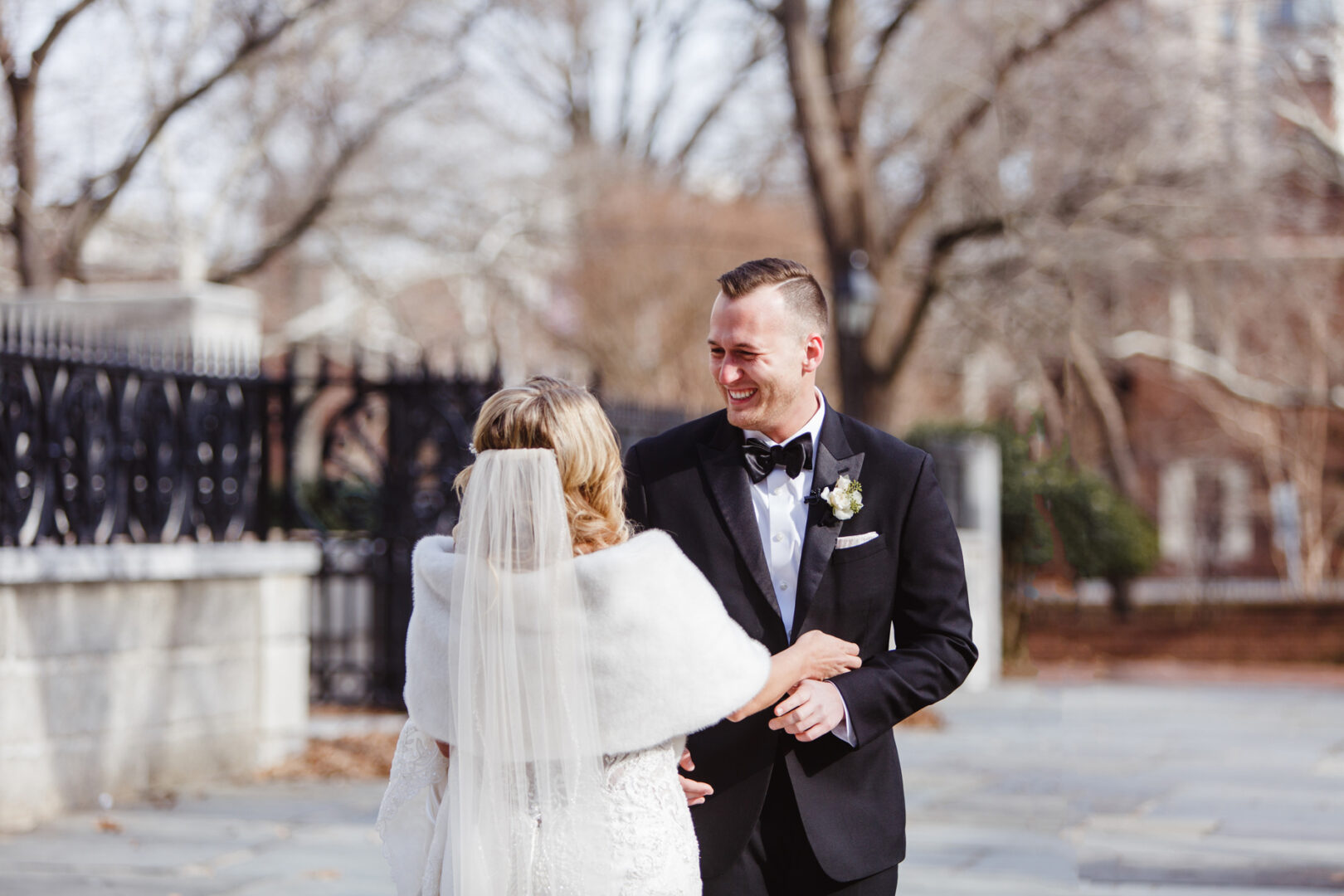 A bride and groom exchange smiles outdoors, with the groom in a black tuxedo and the bride in a white dress and veil. They stand on a stone path, framed by bare trees, echoing the elegance of their Downtown Club wedding.