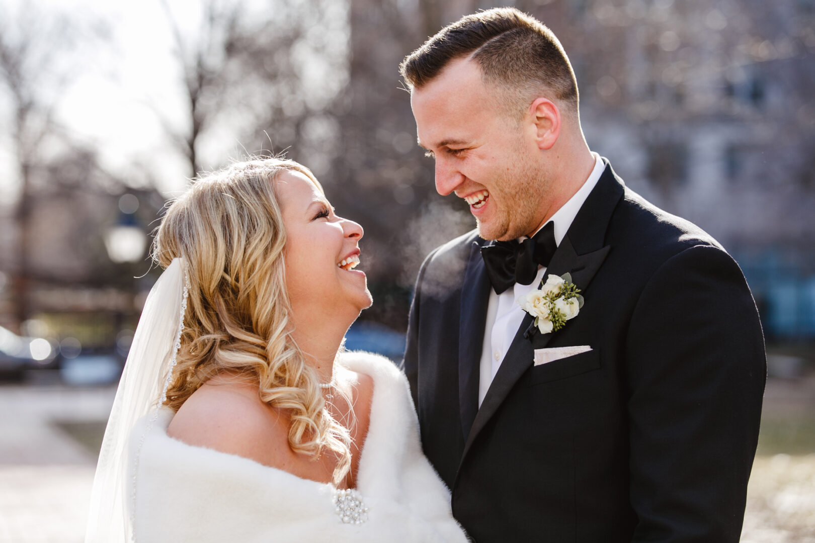 A bride and groom in formal attire laugh and smile at each other outdoors on a sunny day, capturing the joyous spirit of their downtown club wedding.