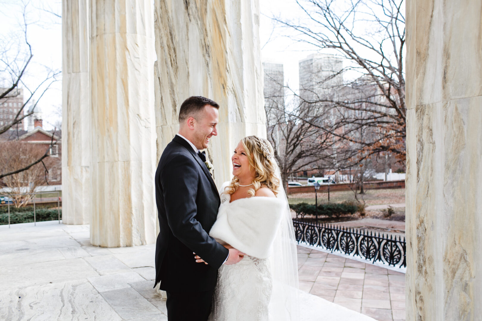 A couple in formal attire, the woman in a white dress with a shawl and the man in a dark suit, stand happily together against the backdrop of large stone columns and trees, celebrating their Downtown Club wedding.