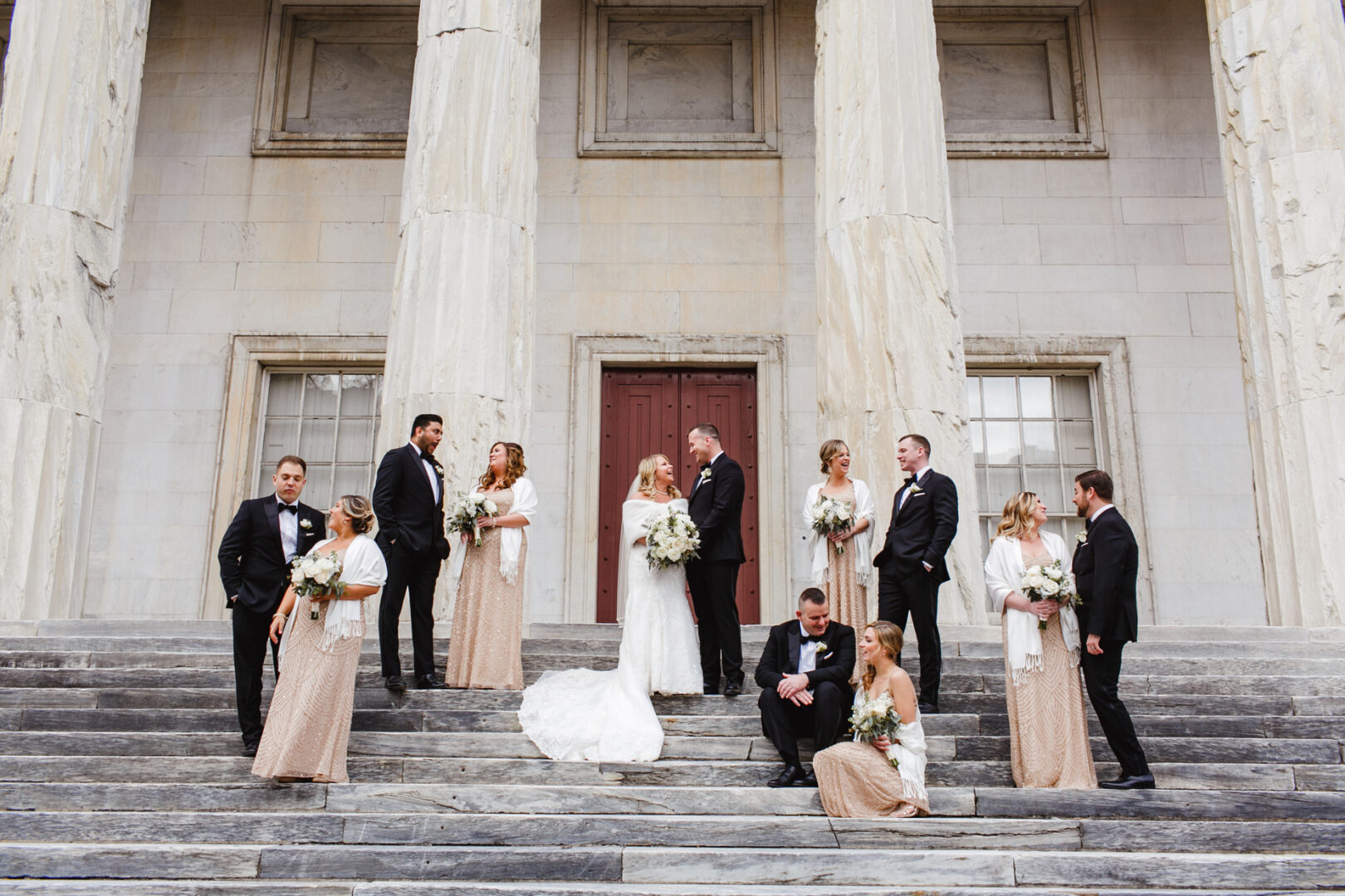The bridal party is elegantly posed on the stone steps of a grand columned building, echoing the charm of a Downtown Club wedding. Bridesmaids hold bouquets while groomsmen in black suits flank the radiant bride and groom at the center.