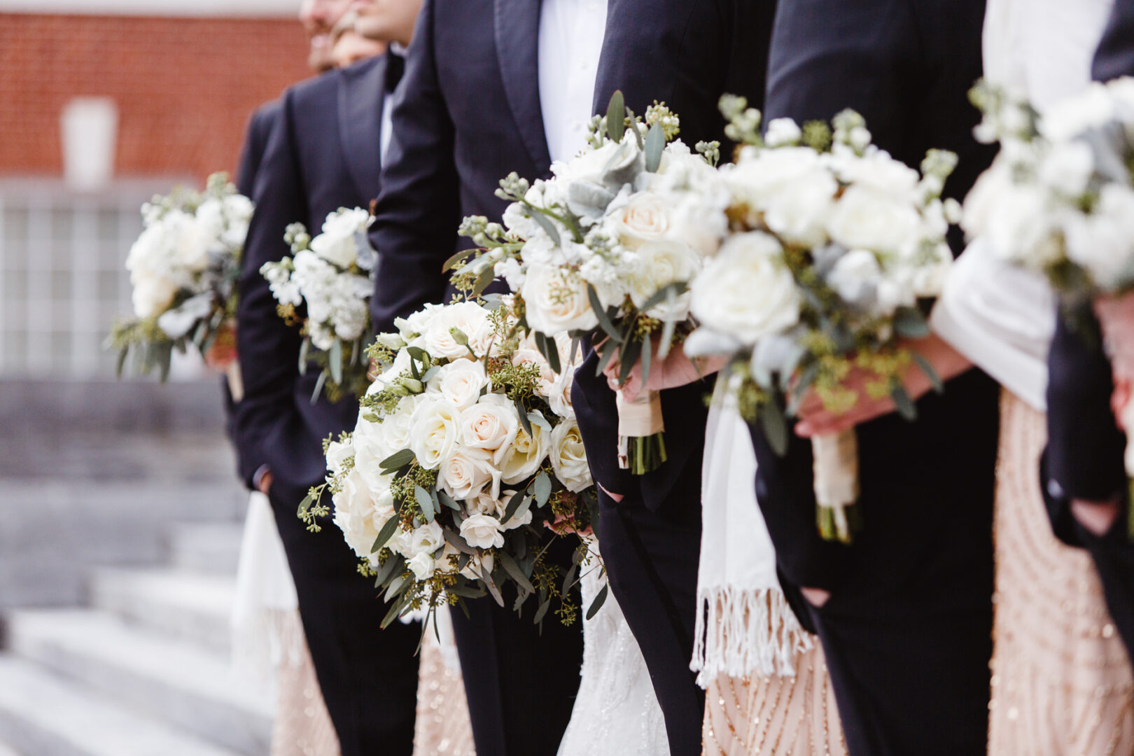 A wedding party stands in line holding bouquets of white and green flowers, with groomsmen in dark suits and bridesmaids in light dresses outside a brick building, perfectly capturing the charm of a Downtown Club wedding.