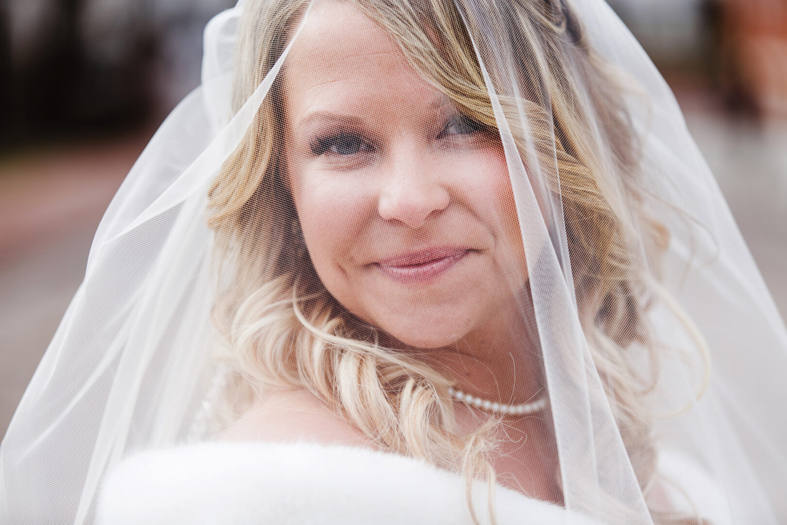 A bride with blonde hair smiles while wearing a white veil and pearl necklace, capturing the elegance of a Downtown Club wedding outdoors.
