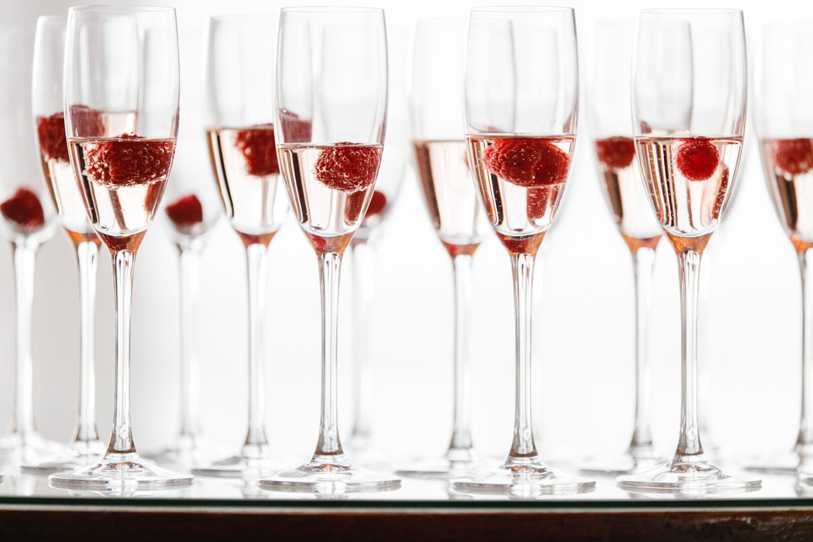At the downtown club wedding, rows of champagne glasses filled with pink sparkling wine gleam elegantly, each adorned with a vibrant red raspberry.