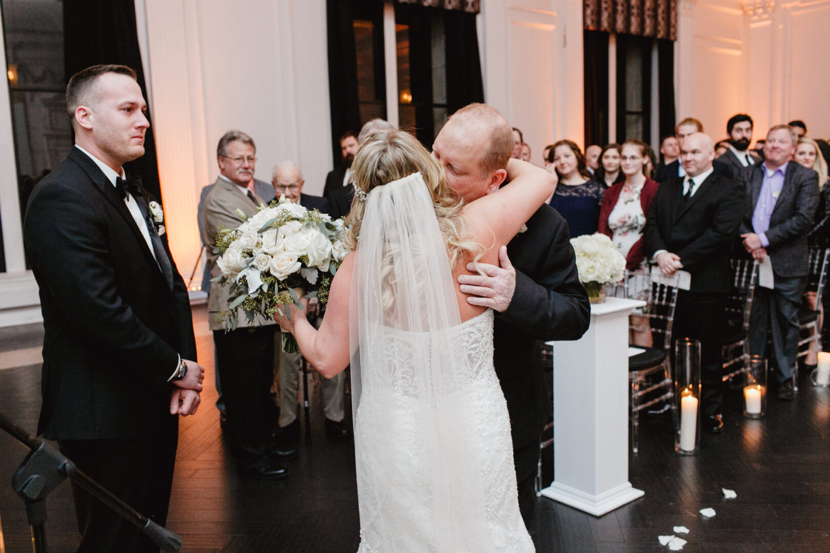 A bride in a white dress embraces an older man during a Downtown Club wedding ceremony, while a man in a black suit stands nearby. Guests watch from their seats in the elegantly decorated venue.