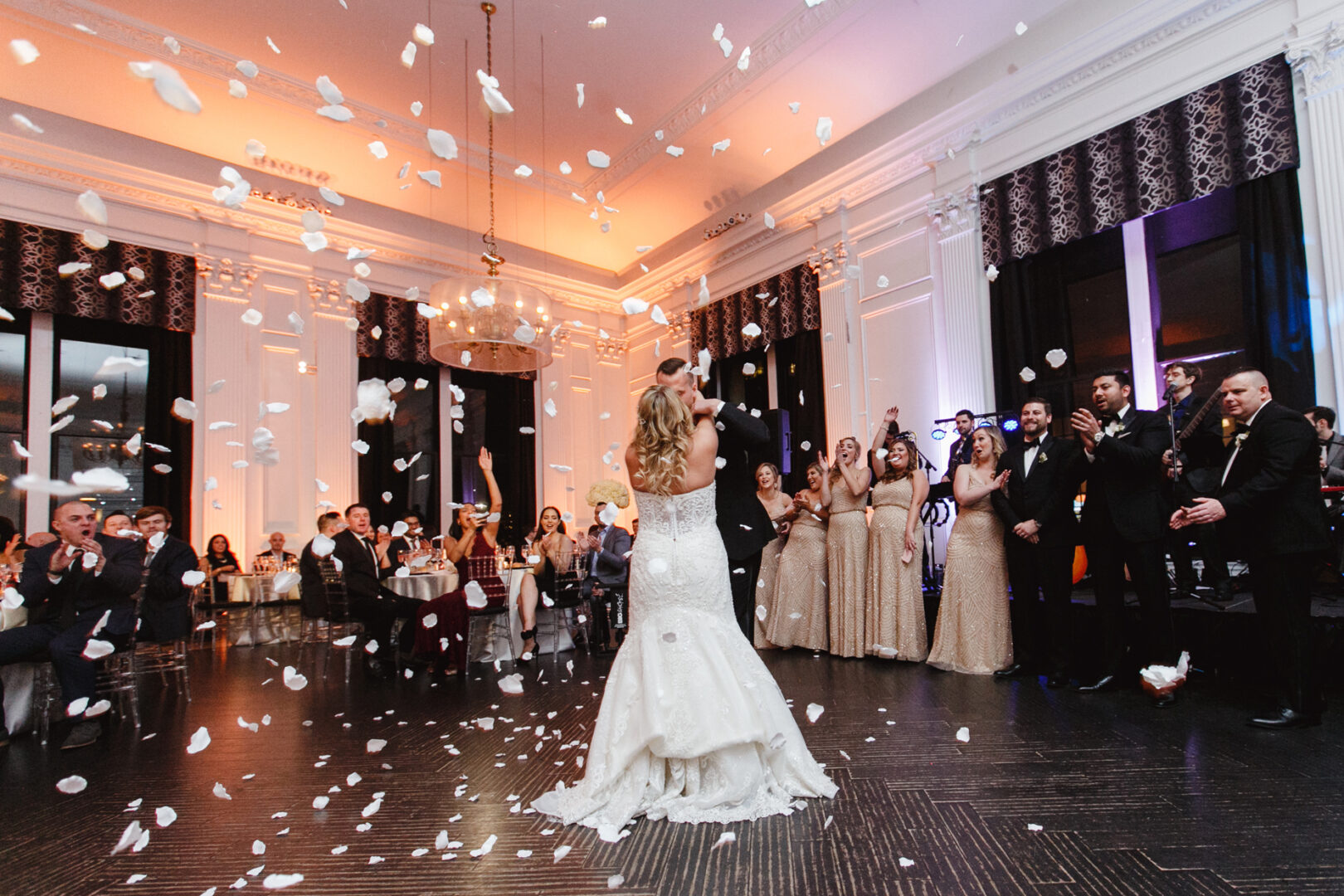 In an elegantly adorned room at a downtown club wedding, a bride tosses her bouquet into the crowd as onlookers cheer and flower petals gently fall around them.