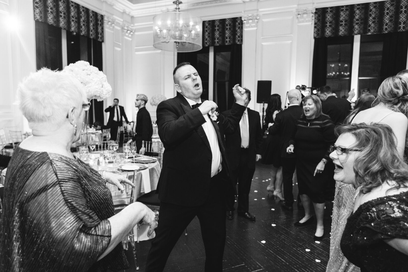 At a downtown club wedding, a man in a suit enthusiastically dances amidst the elegantly dressed crowd, all gathered in a large, beautifully decorated room.