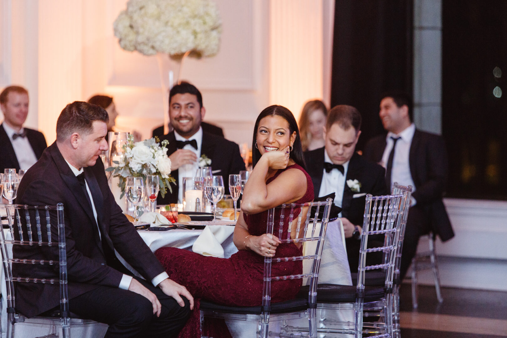 Guests seated at a Downtown Club wedding, with tables set for dining. One woman in a burgundy dress is smiling. The elegant setting includes floral arrangements and luxurious tableware, creating an atmosphere of refined celebration.