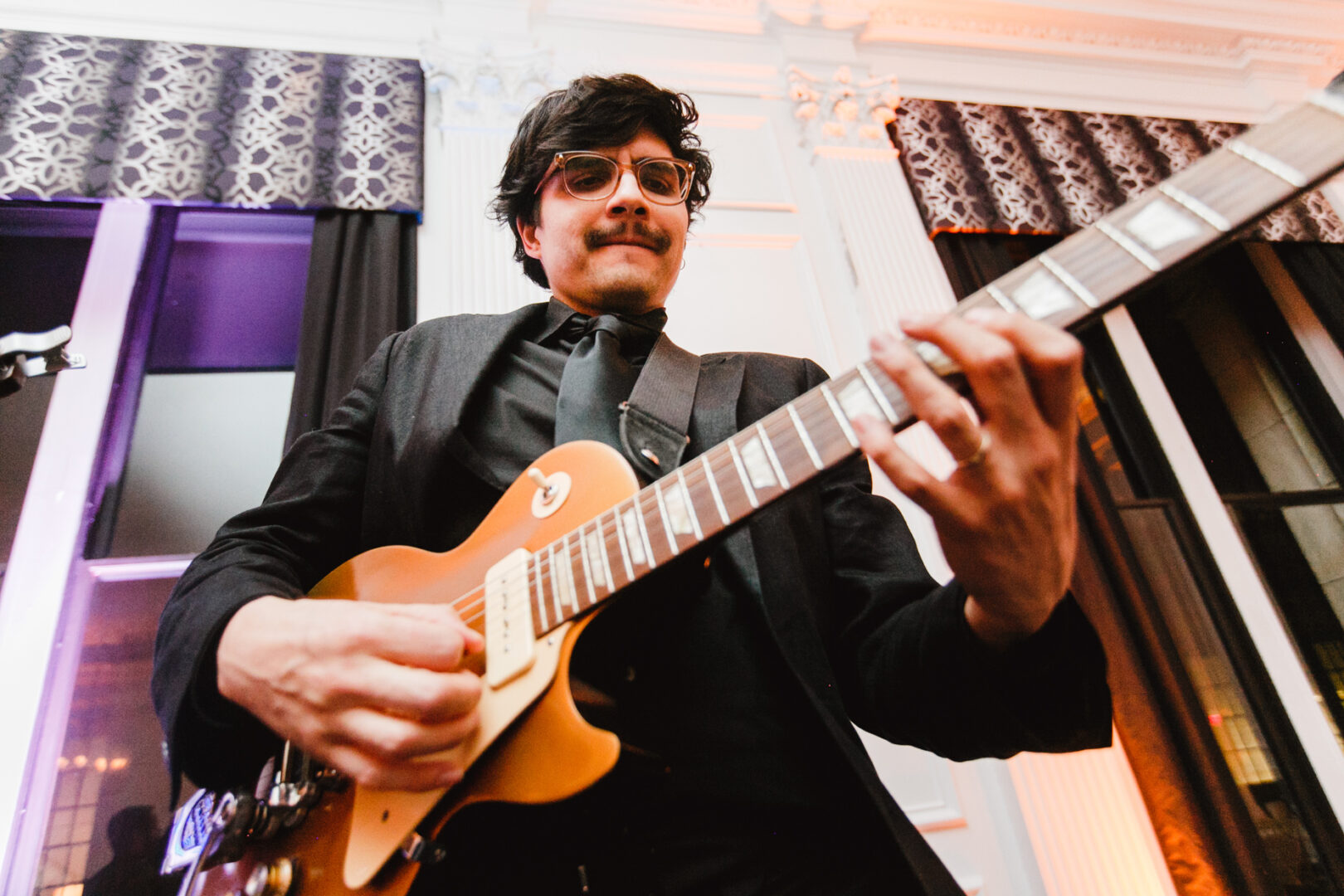 A musician with glasses playing an electric guitar, wearing a dark suit, stands in front of a patterned curtain in a well-lit downtown club.