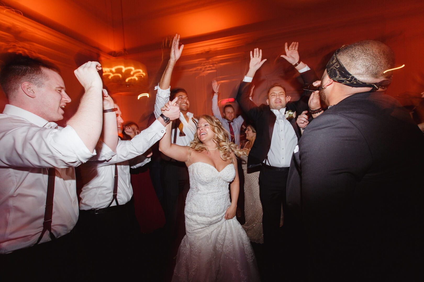 At a vibrant downtown club wedding, people are dancing energetically at the reception. The bride, glowing in her white dress, commands the center as warm red-toned lights set the celebratory mood.