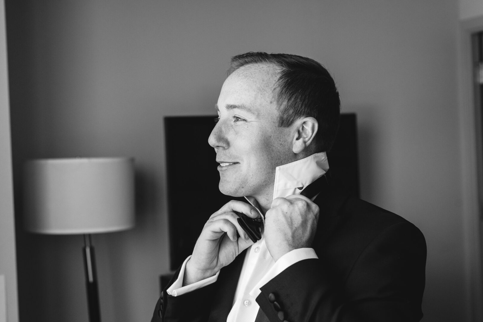 A man in a suit adjusts his collar beside a lamp, capturing the timeless elegance reminiscent of a Penn Museum wedding. The black and white photo exudes classic charm.