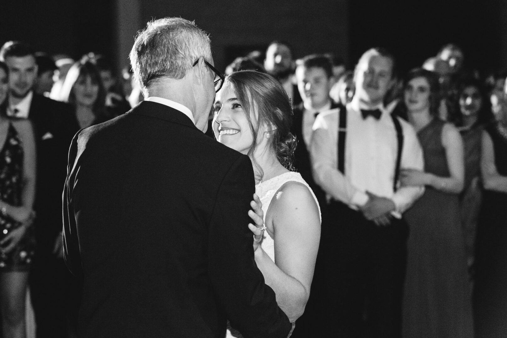 At a Penn Museum wedding, a woman in a white dress gracefully dances with a man in a suit. They are surrounded by an elegantly attired crowd, captivated by their performance.
