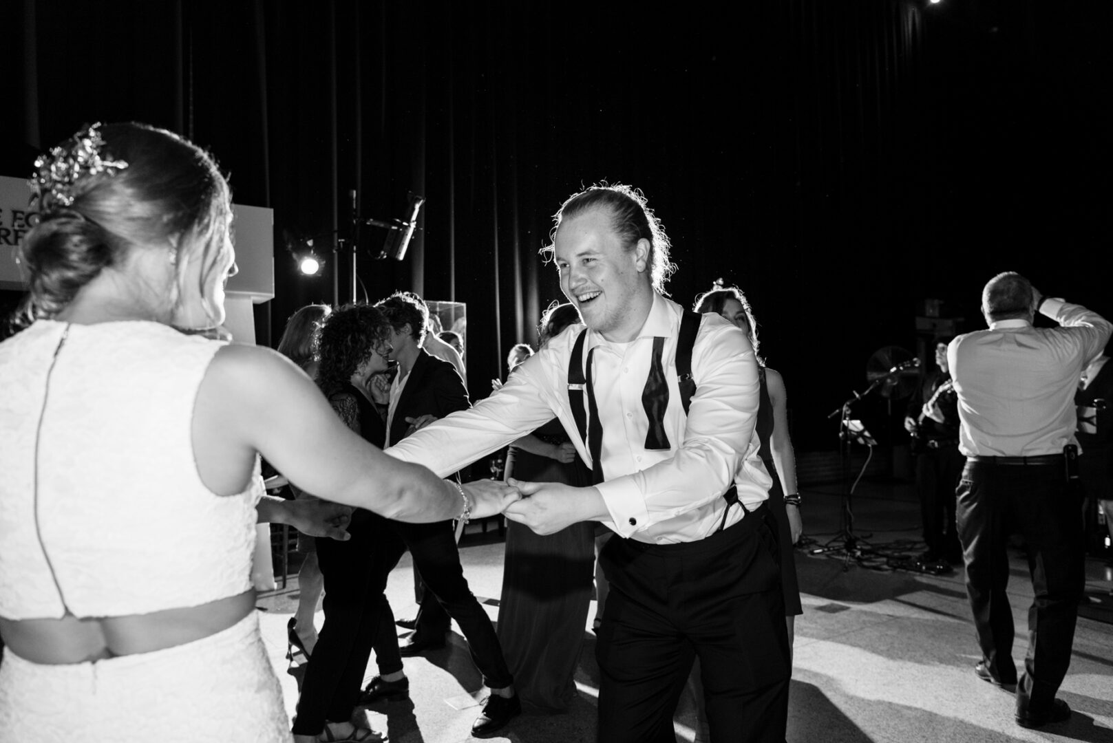 At a Penn Museum wedding, a couple dances gracefully amidst the ancient artifacts. The man, in a dress shirt and suspenders, guides his partner in a sleeveless dress, while others look on, immersed in this beautiful blend of history and romance.