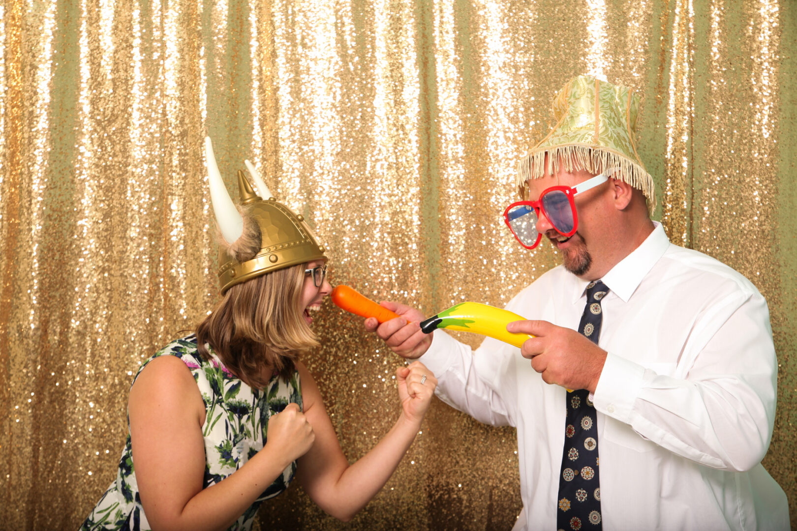Two people, donning fun hats and oversized glasses, jovially pretend to duel with a plastic banana and carrot against a gold sequin backdrop in the Cape May Photo Booth's whimsical atmosphere.