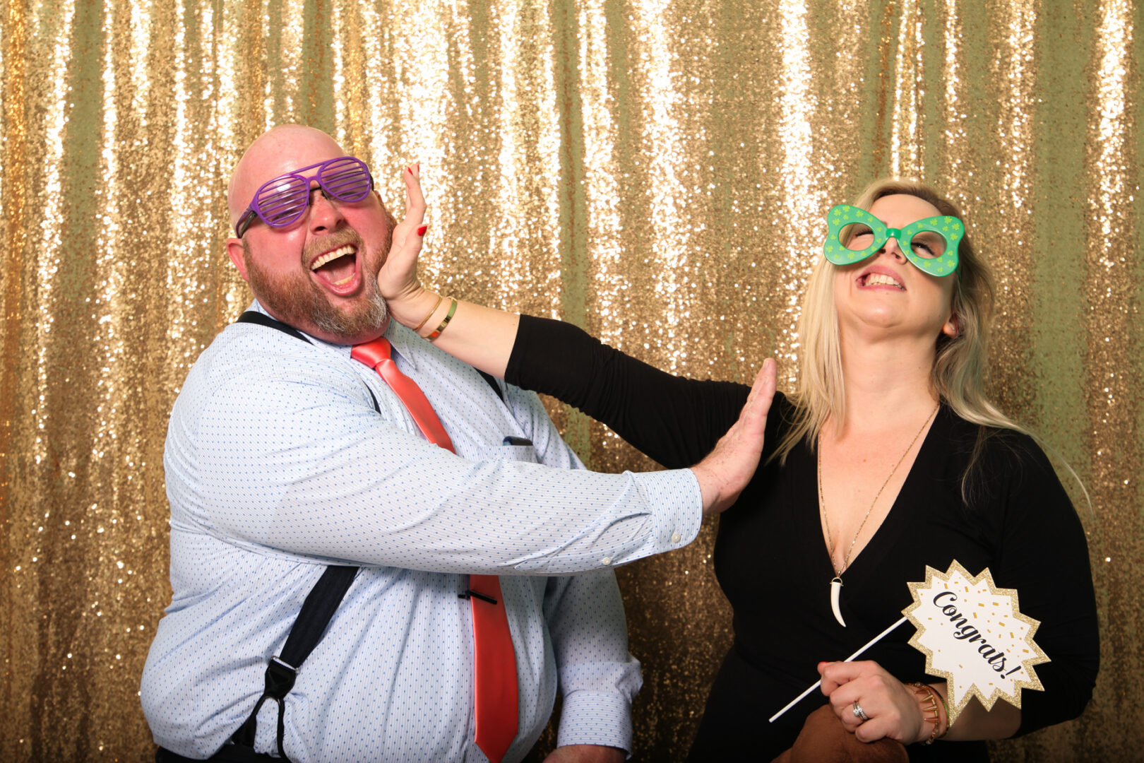 Two people laughing and posing playfully with oversized glasses in front of a gold sequin backdrop from Cape May Photo Booth. One holds a "Congrats" sign, adding to the fun atmosphere.