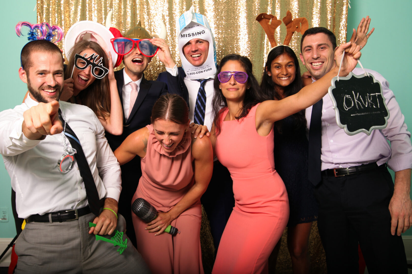 A group of people posing playfully in front of a Cape May Photo Booth's gold sequin backdrop, wearing various props like hats and glasses, with some holding signs.