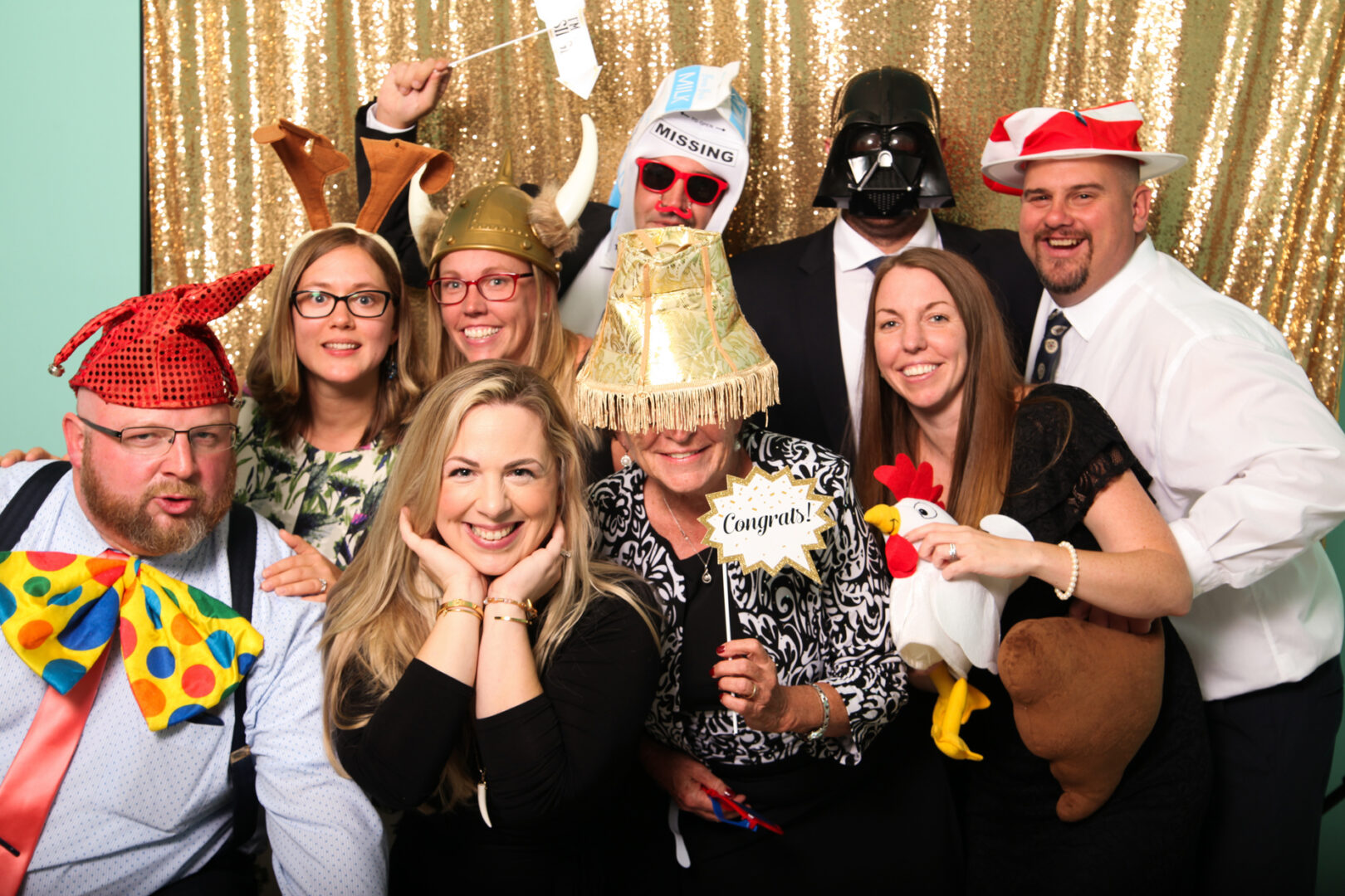 A lively group, decked out in costumes and accessories like hats, masks, and props—complete with a chicken and large bow tie—strike playful poses against a gold sequin backdrop. Their infectious energy makes this Cape May Photo Booth experience unforgettable.