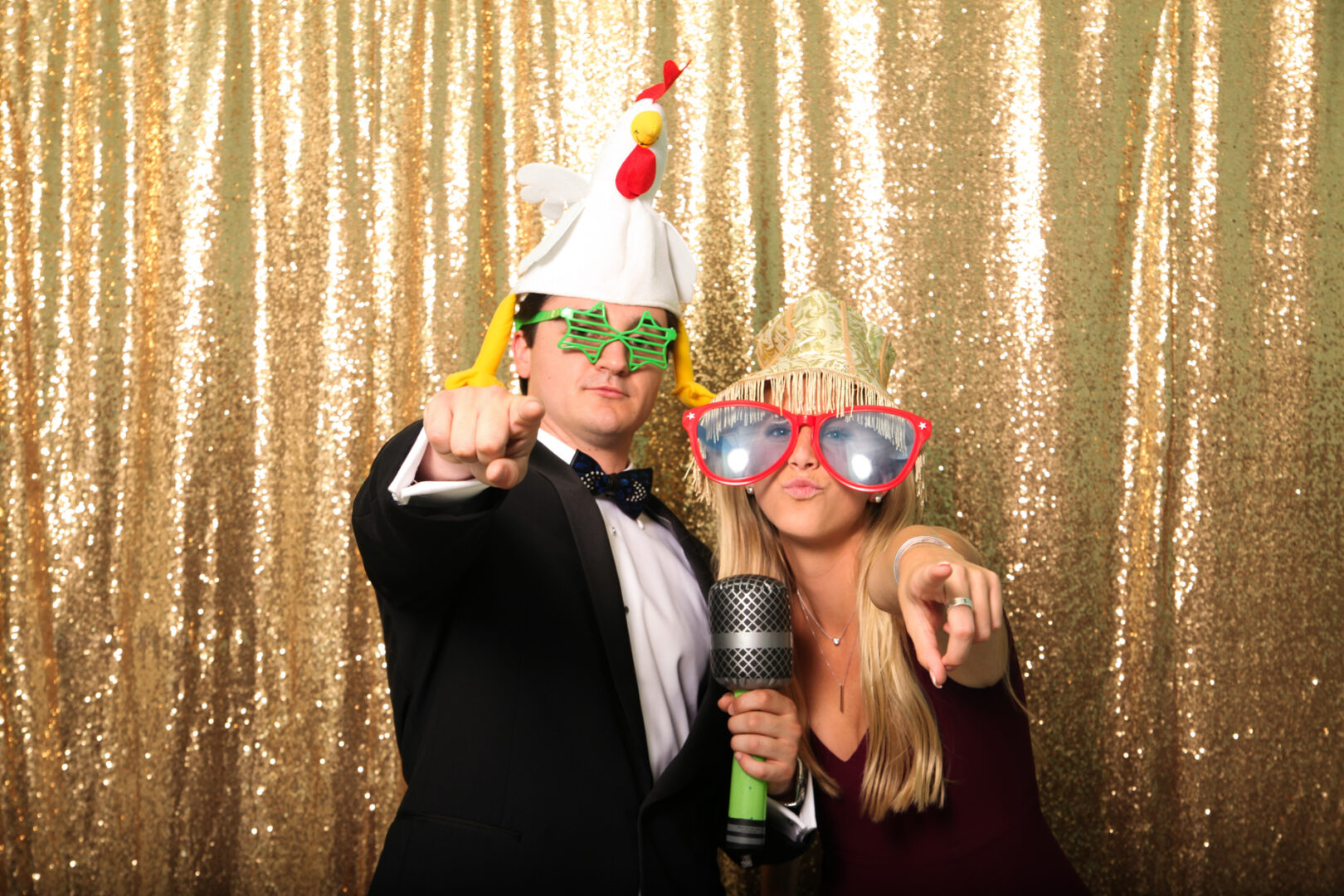 Two people pose playfully in front of a Cape May Photo Booth's sparkling gold sequin backdrop, adorned with novelty hats and oversized glasses. They hold a toy microphone, each pointing towards the camera with infectious joy.