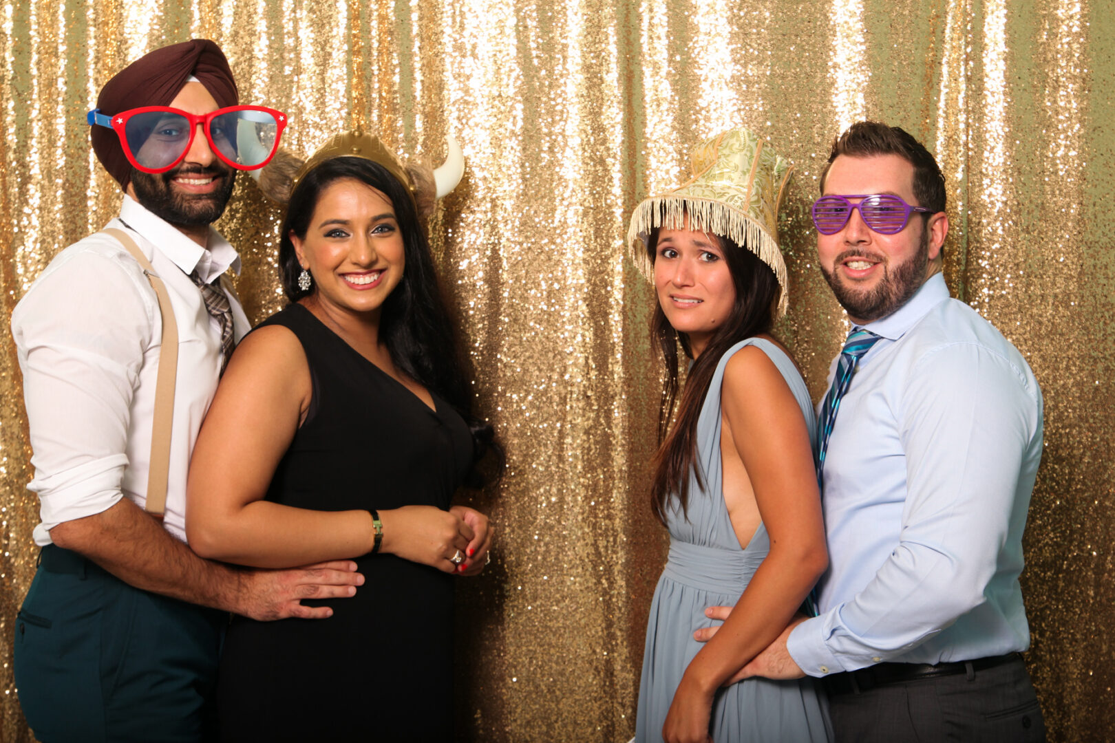 Four people, decked out in playful accessories, smile at the camera in front of a glittering gold backdrop, capturing a fun moment with Cape May Photo Booth.