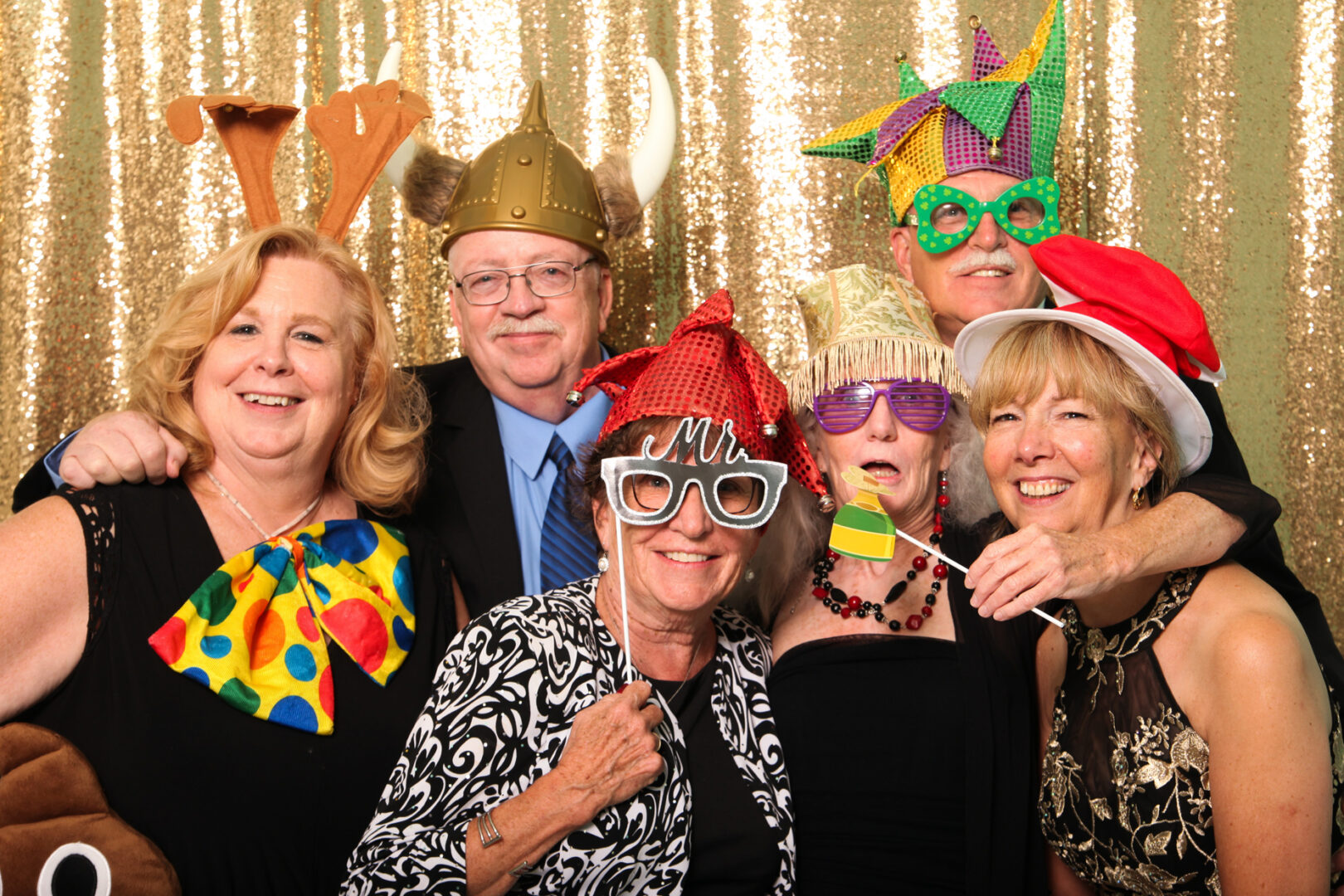 Six people in festive attire and playful hats pose against a gold sequin backdrop, smiling and wearing various novelty accessories, capturing the joyful spirit of a Cape May Photo Booth experience.