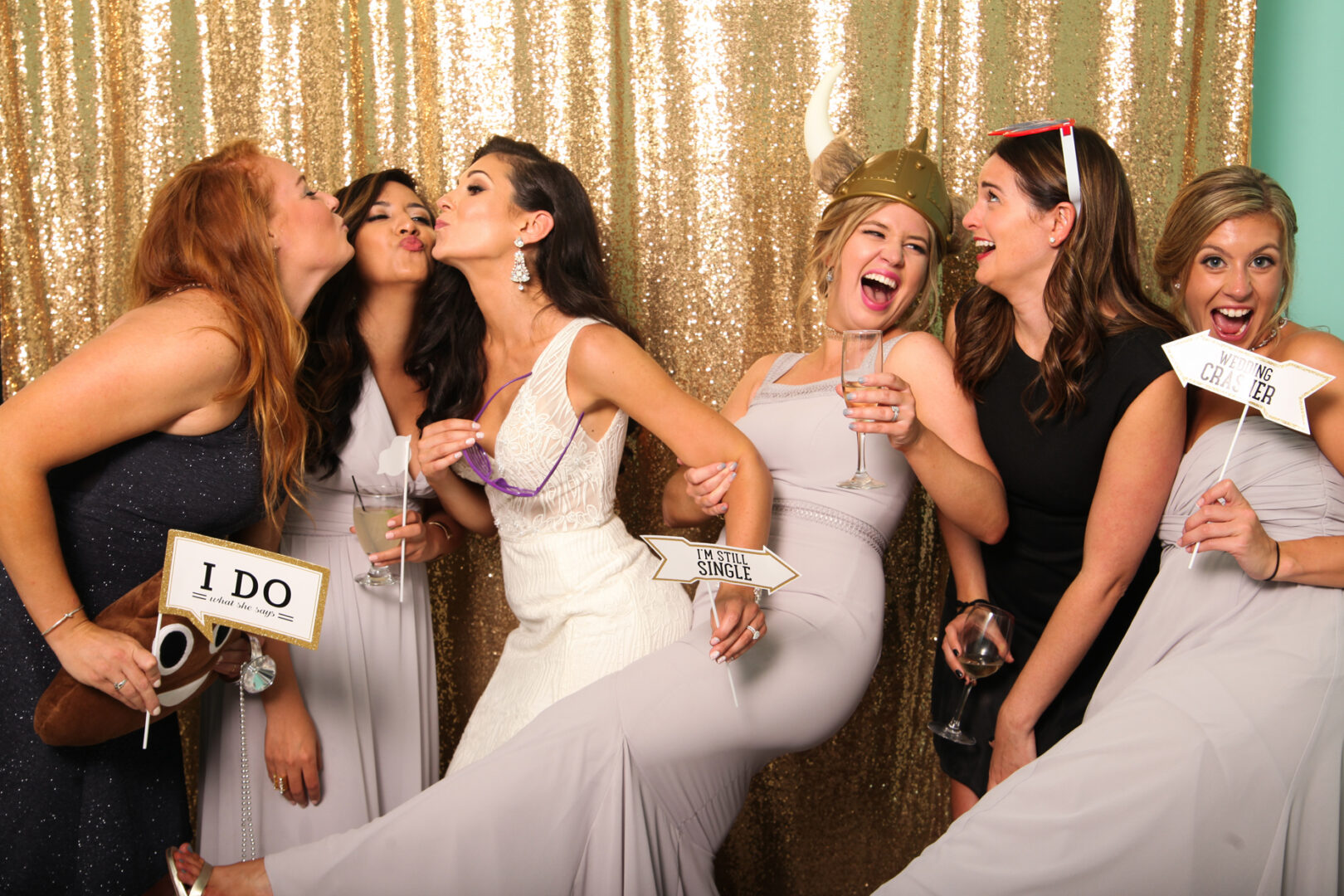 Six women in formal attire strike a playful pose in front of a Cape May Photo Booth’s gold sequin backdrop, holding signs like "I DO" and "Single." One even sports a Viking hat, as they smile and laugh together.