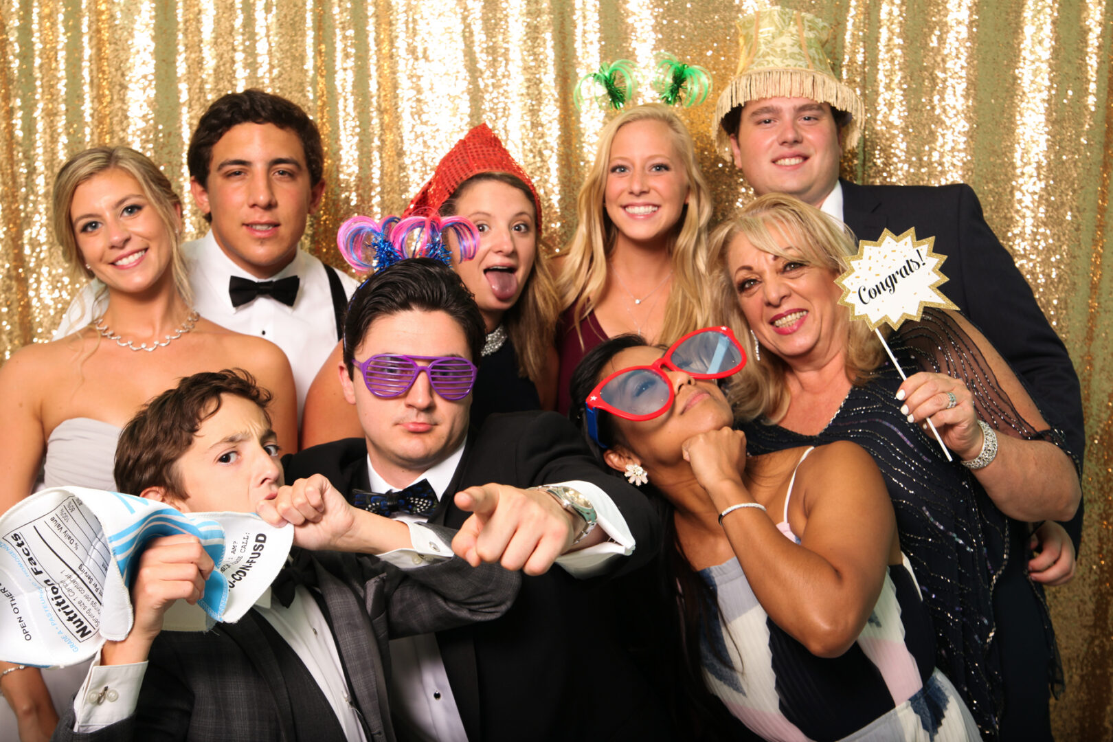 A lively group, dressed in formal attire and party accessories, poses in front of a shimmering gold sequin backdrop. With hats, glasses, and props in hand, the Cape May Photo Booth captures their joyous spirits with a touch of glamour and fun.