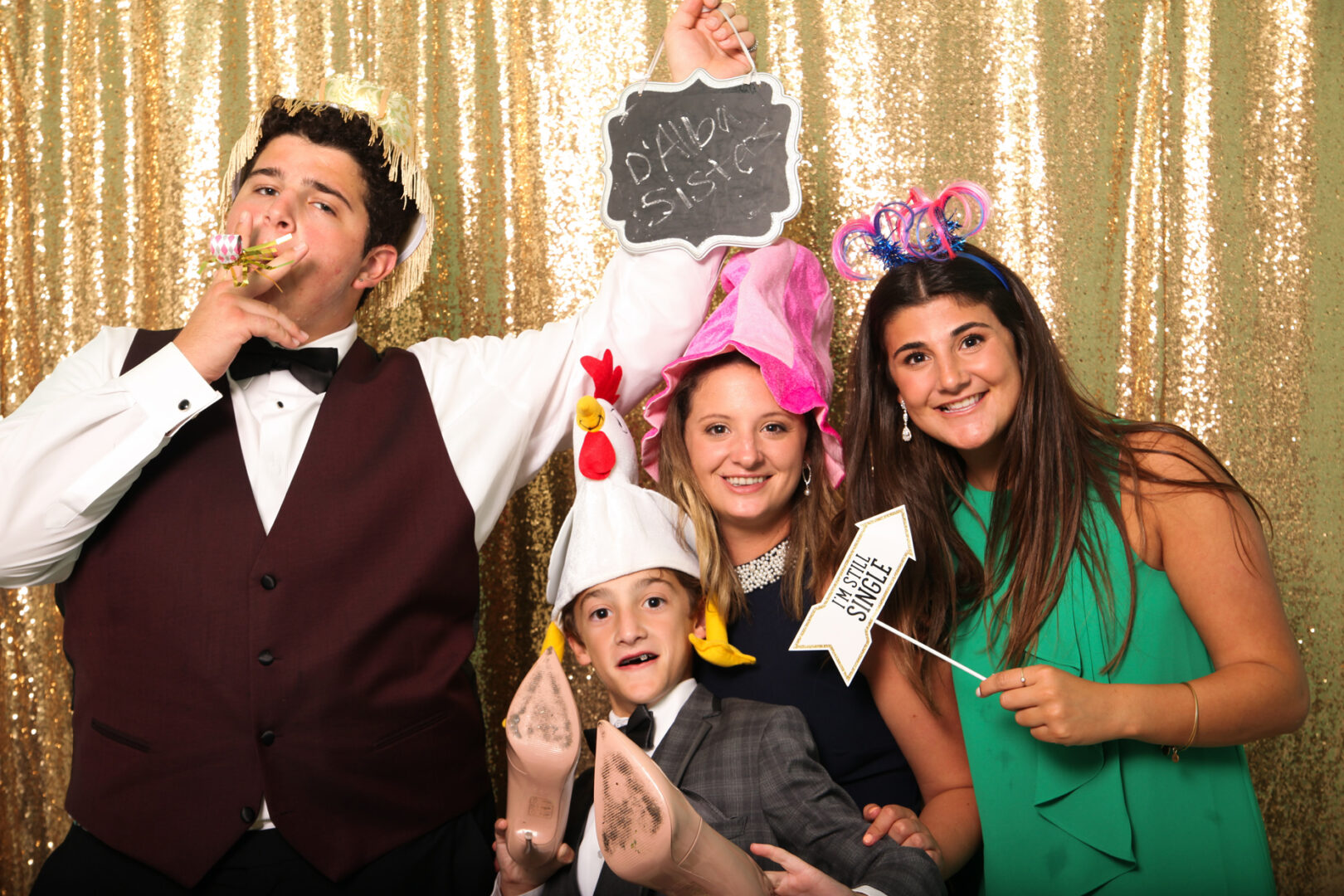 Four people stand before a gold backdrop at Cape May Photo Booth, adorned in playful hats and holding quirky props. One brandishes a sign reading "Single," while another merrily blows a party horn, capturing the joyous spirit of the moment.