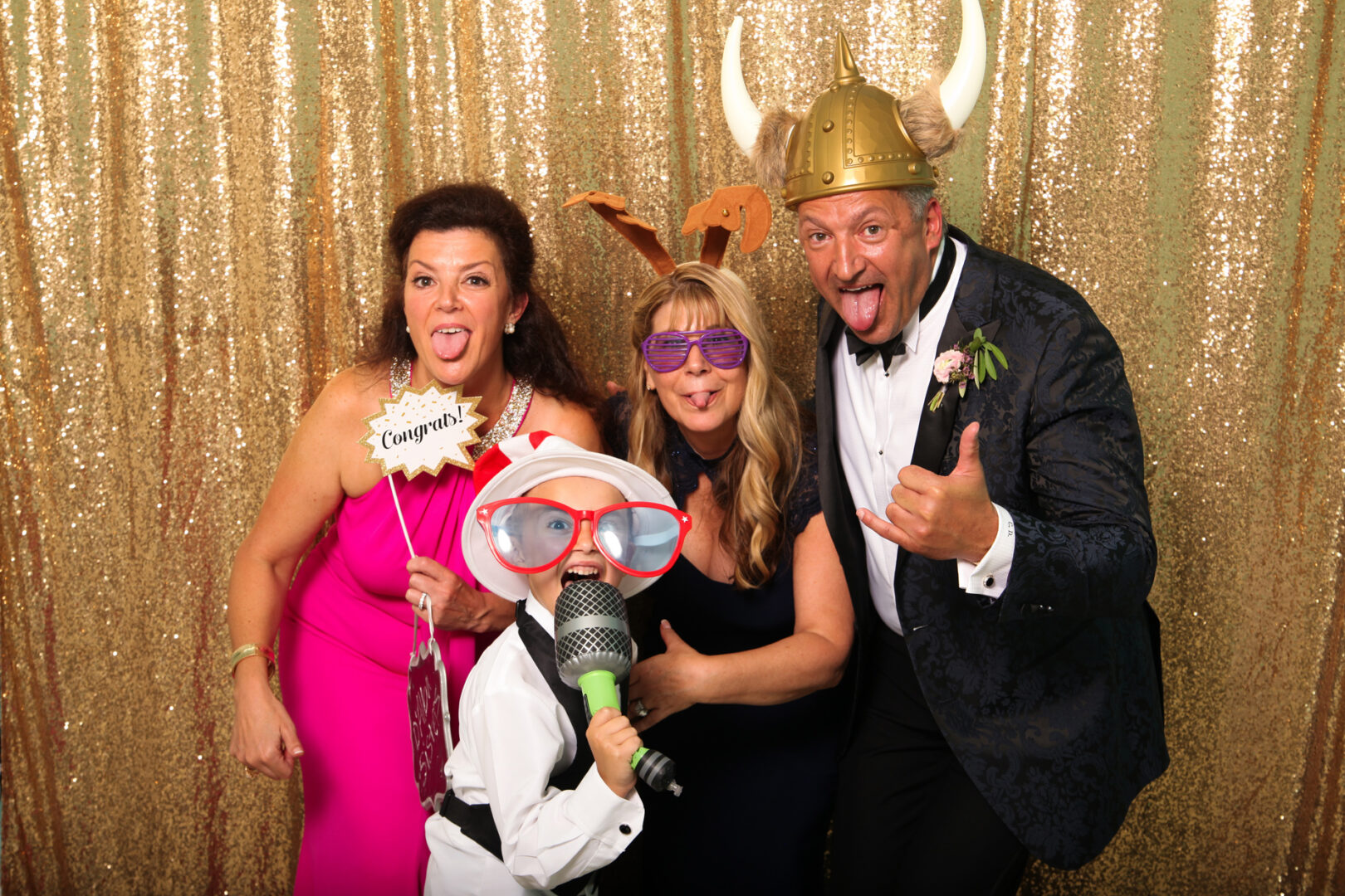Four people pose playfully in front of a gold sequin backdrop at the Cape May Photo Booth. They wear novelty hats, glasses, and hold props, smiling and sticking out their tongues.