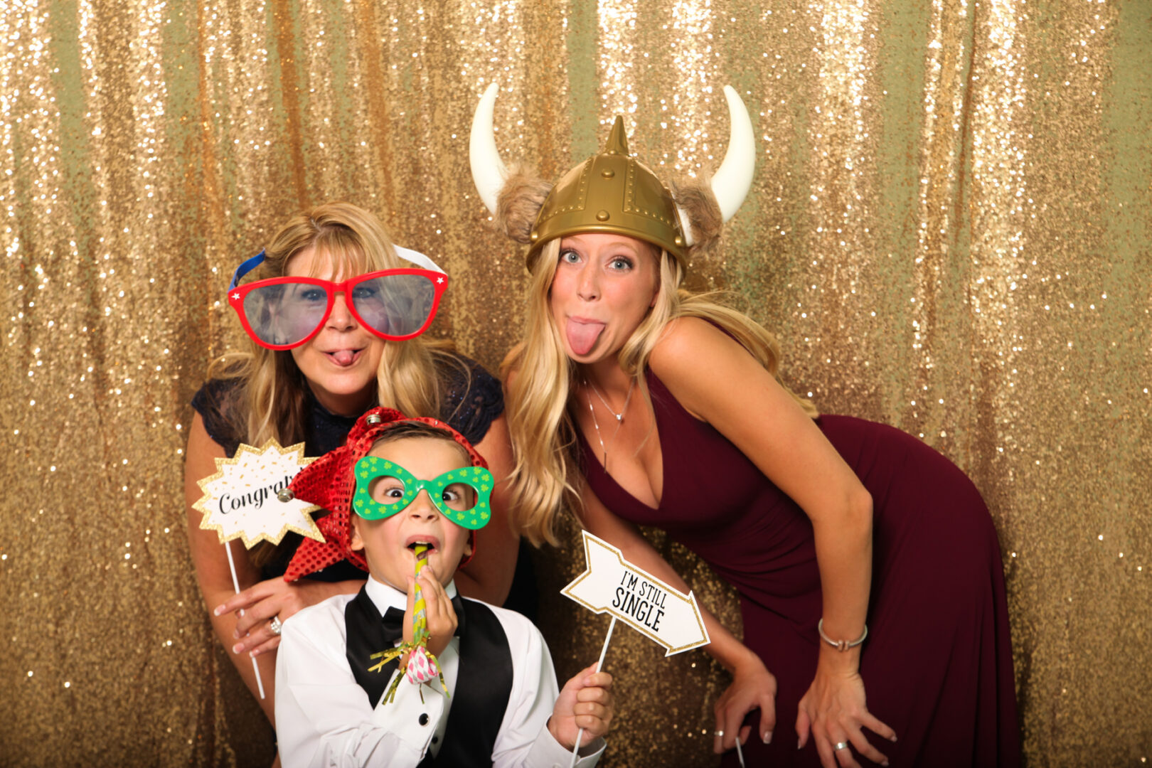 Three people posing playfully in front of a gold sequin backdrop at the Cape May Photo Booth. They wear oversized glasses and novelty hats, holding humorous signs.