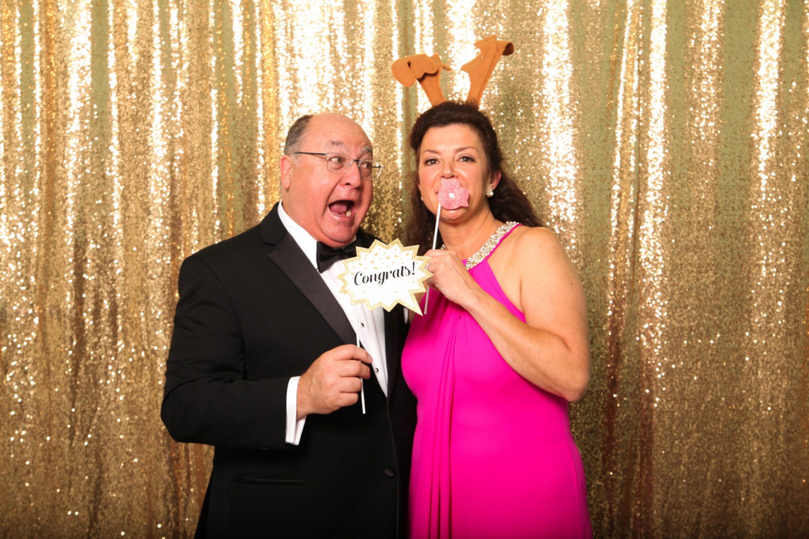 A man in a tuxedo and a woman in a pink dress with an antler headband strike playful poses with fun props against a stunning Cape May Photo Booth gold sequin backdrop.